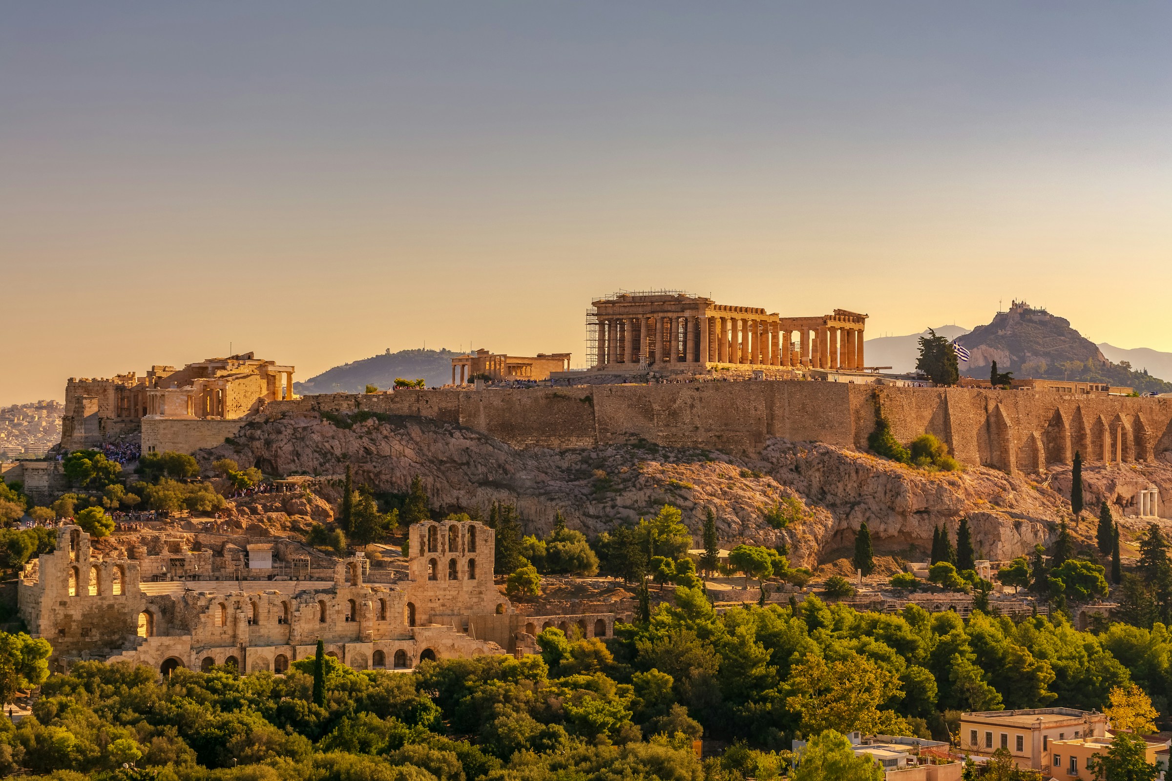 View of the Acropolis of Athens at sunset, featuring the Parthenon and surrounding ancient structures, with lush greenery in the foreground.
