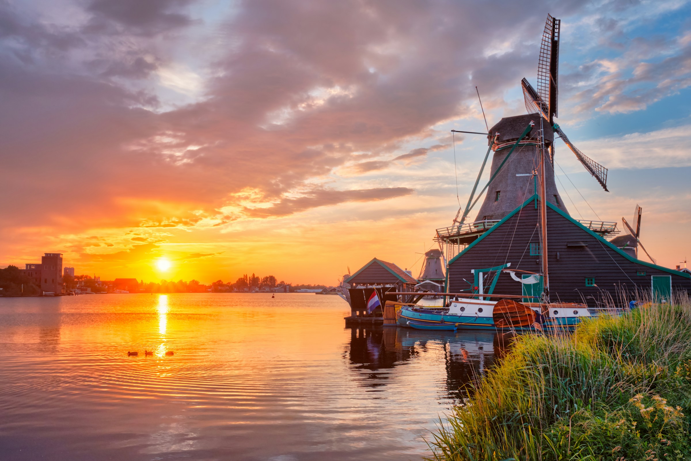 View of a traditional Dutch windmill near a lake during sunset, with vibrant orange and pink skies reflecting on the water.