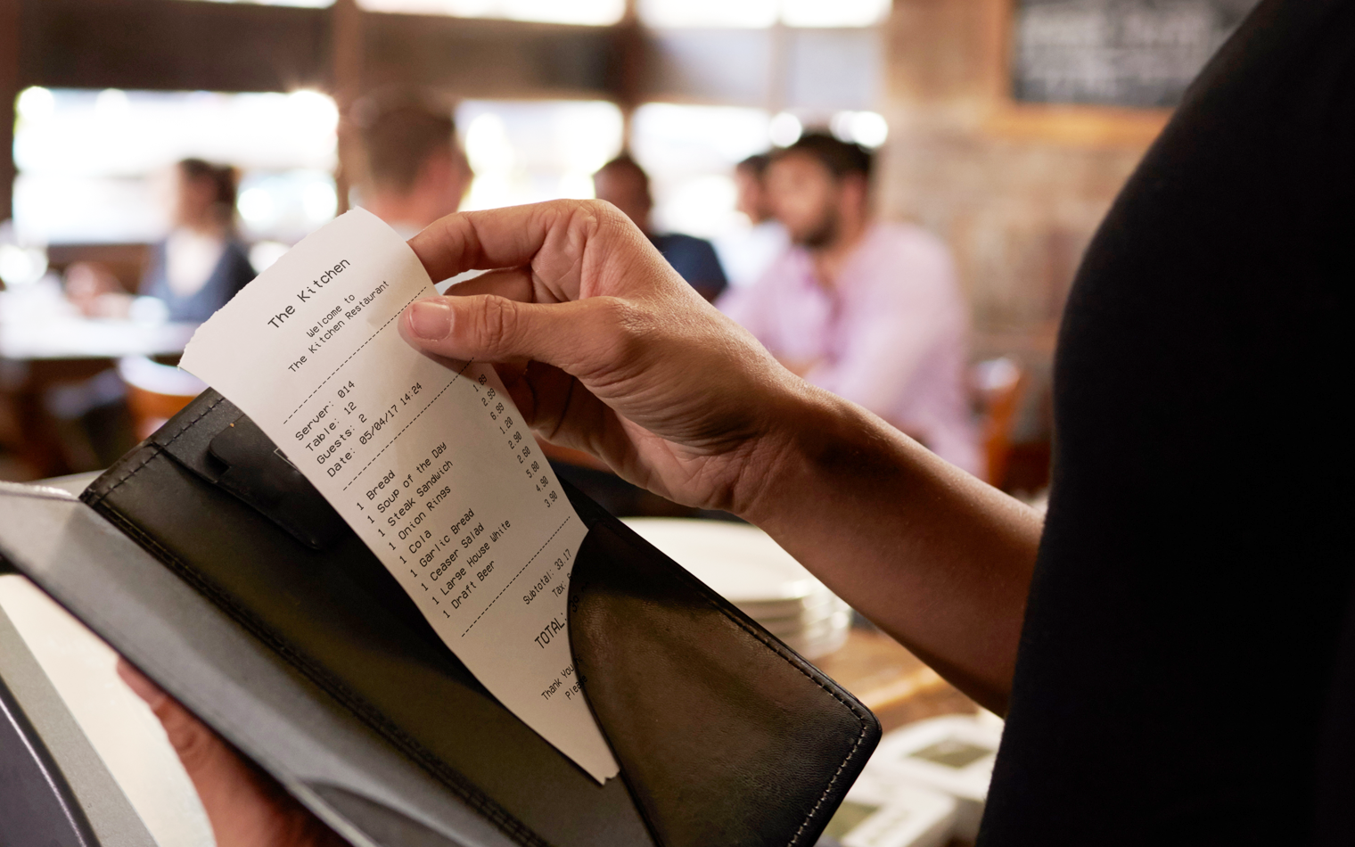 Henkilö, joka pitää ravintolalaskua nahkapidikkeessä istuessaan pöydän ääressä ravintolassa. Taustalla näkyy useita ihmisiä, samoin kuin sumea ravintolan sisätila.