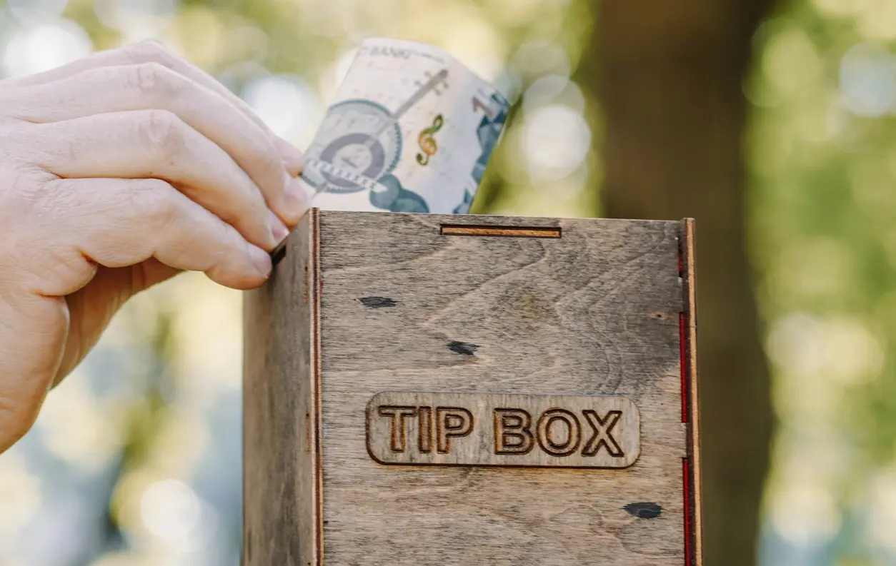 Hand l&auml;gger en sedel i en tr&auml;box m&auml;rkt &ldquo;Tip Box&rdquo;, avsedd f&ouml;r dricks.