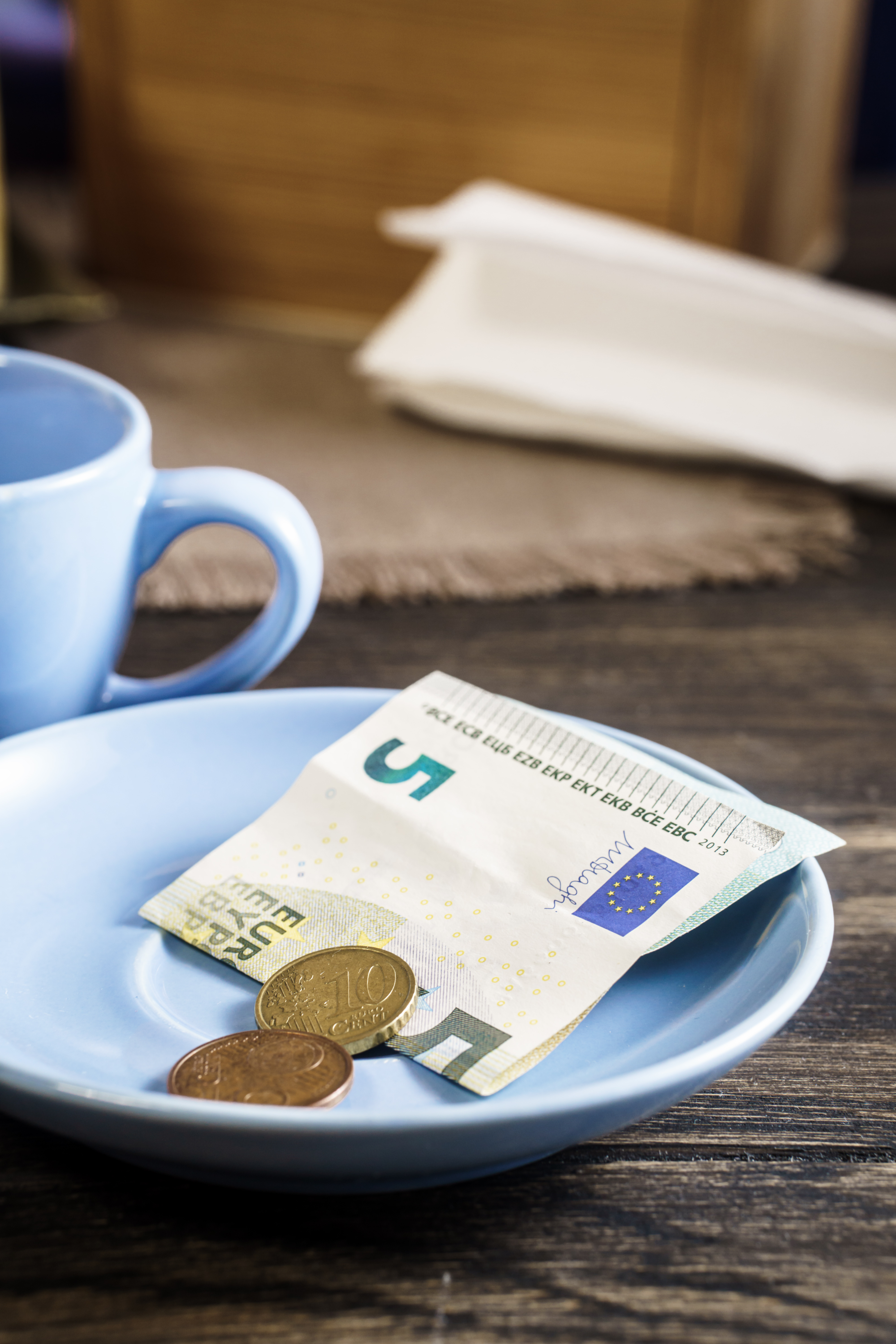 A light blue plate with a five-euro note and coins placed on a wooden table next to a blue coffee cup, creating a cozy café setting.