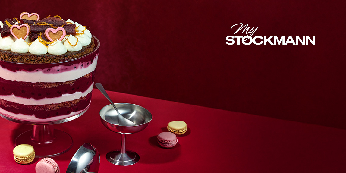 Jälkiruokamalja kerroksittain lakritsaa ja kermaa, sydänkoristeita päällä, punaisella pöydällä macarons-leivosten kanssa, vieressä pieni kastikekulho. Taustalla My Stockmann -logo.