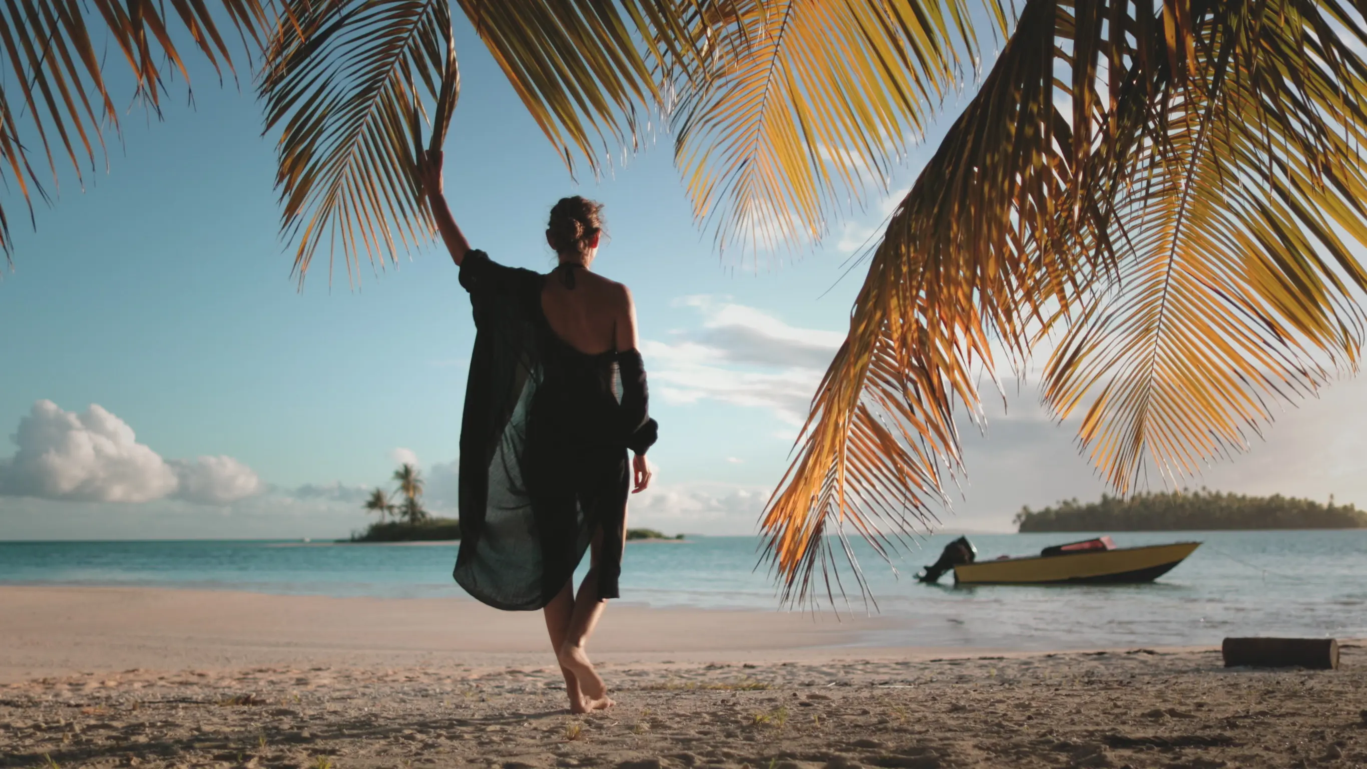 Woman in a black dress walking on a tropical beach under palm trees, with a boat on the ocean and an island in the distance.