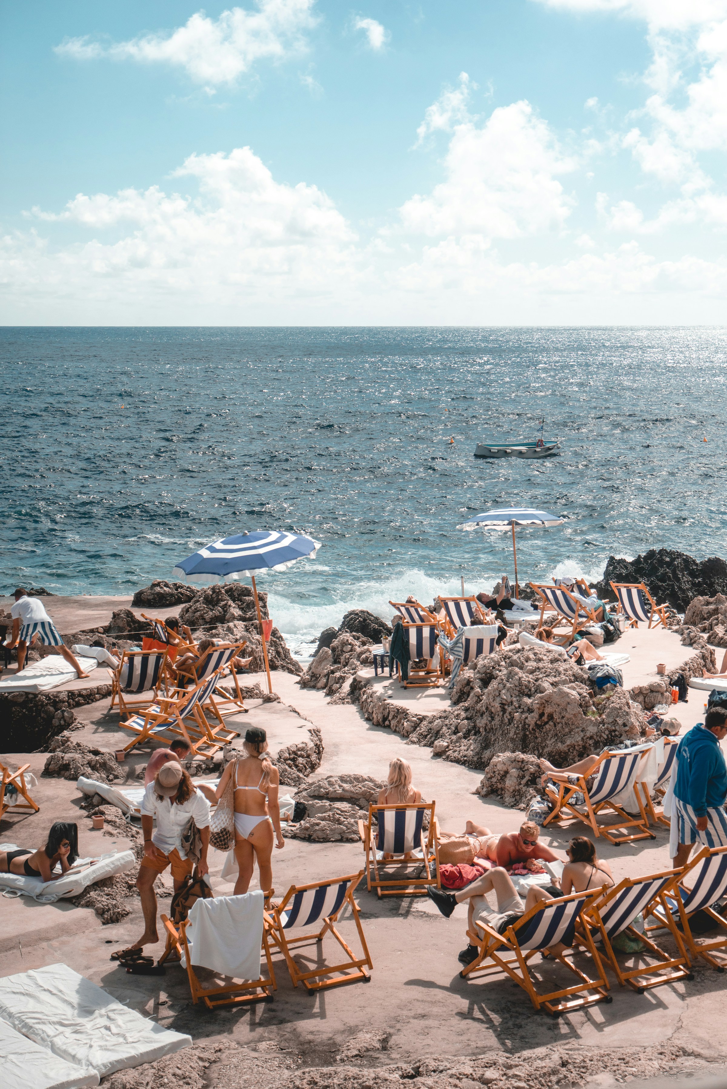 Sun loungers and umbrellas on the rocky coast with turquoise sea and bathers enjoying a beach holiday by the Mediterranean. Relaxing beach environment in Southern Europe.