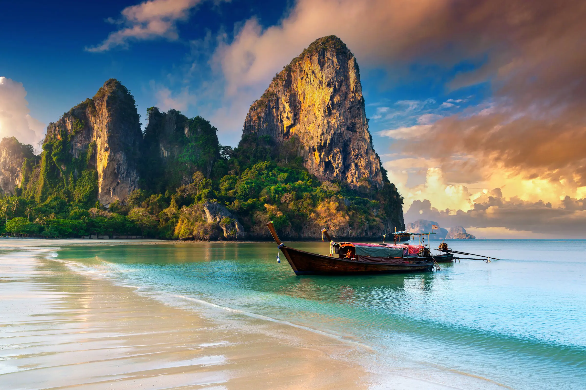 Bästa resorna vintern 2024 - Strand med traditionell thailändsk långbåt, kristallklart vatten och dramatiska kalkstensklippor vid solnedgången i Krabi, Thailand.