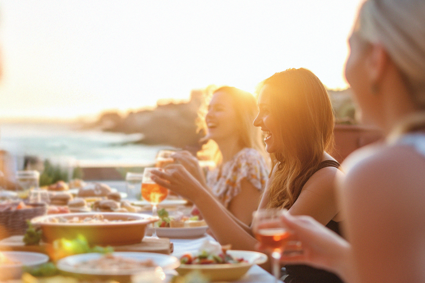 Folk njuter av middag utomhus i solnedgången, skrattar och håller dricksglas i händerna.