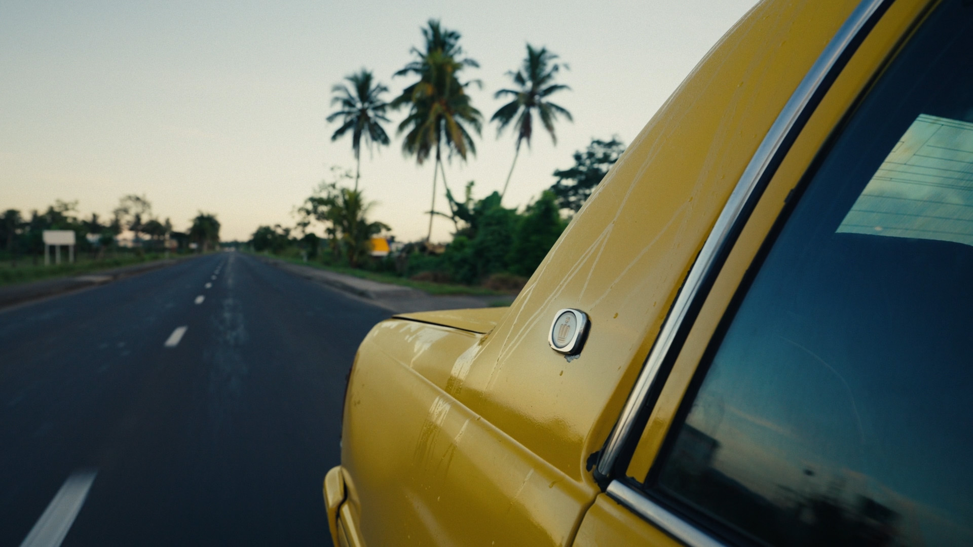 Yellow car driving on a tropical road lined with palm trees under a clear sky.