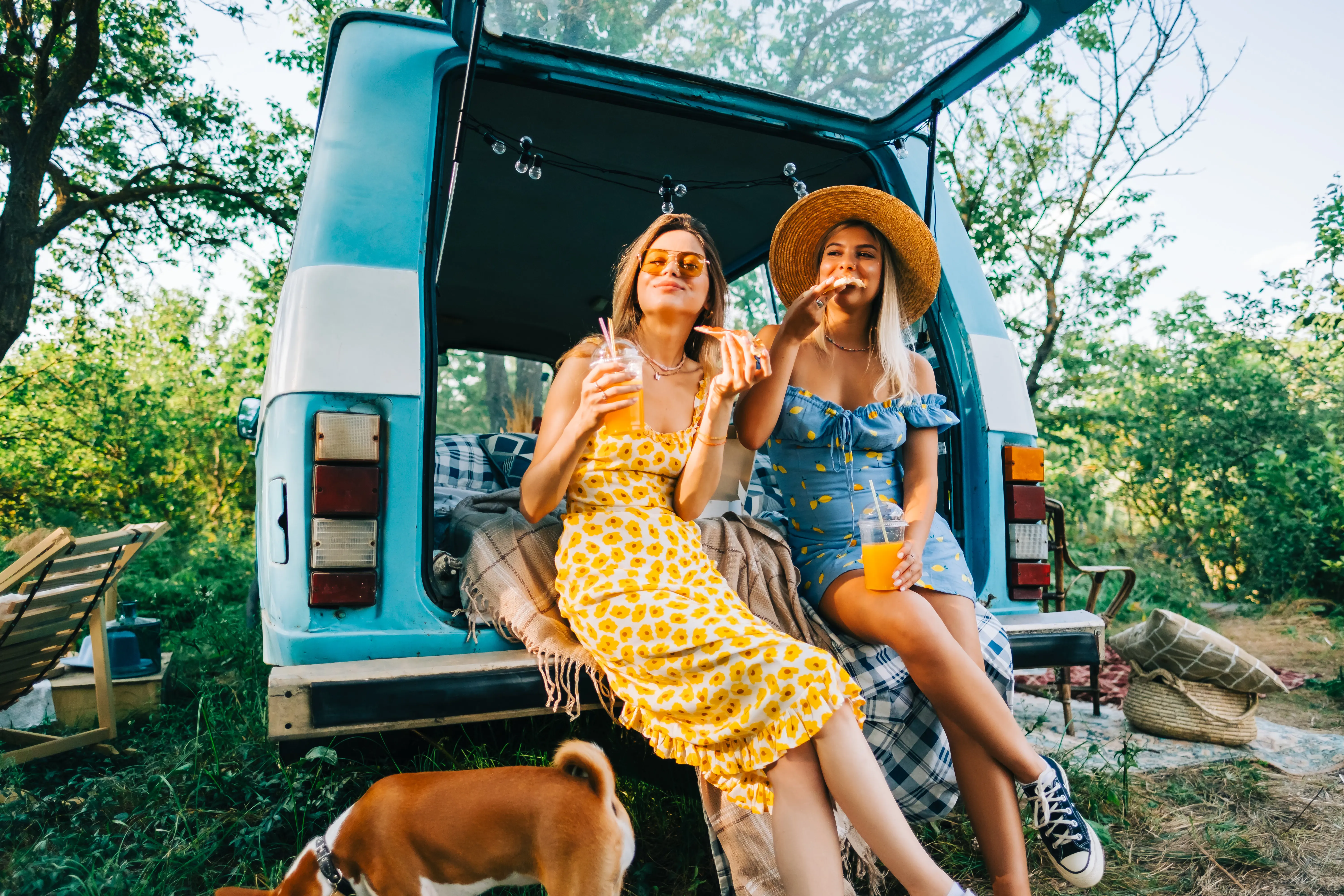 Two women enjoying a summer's day at a campsite, sitting in an open trunk of a retro van with a dog walking around.