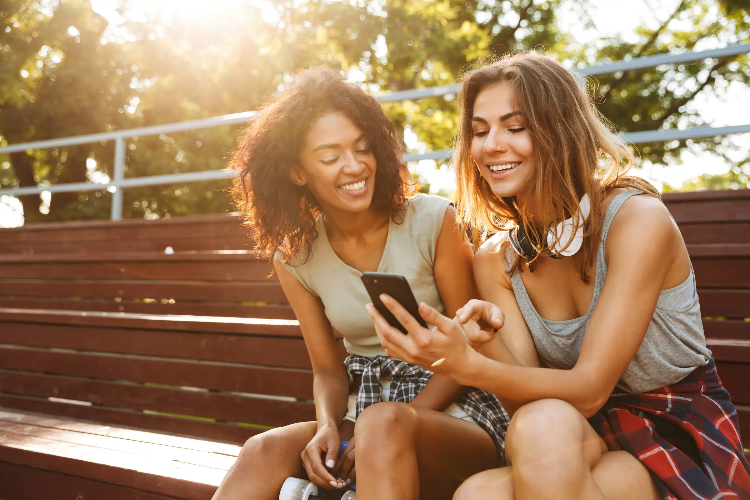 Two young women sitting on wooden bleachers, smiling while looking at a smartphone on a sunny day, wearing casual summer clothing.