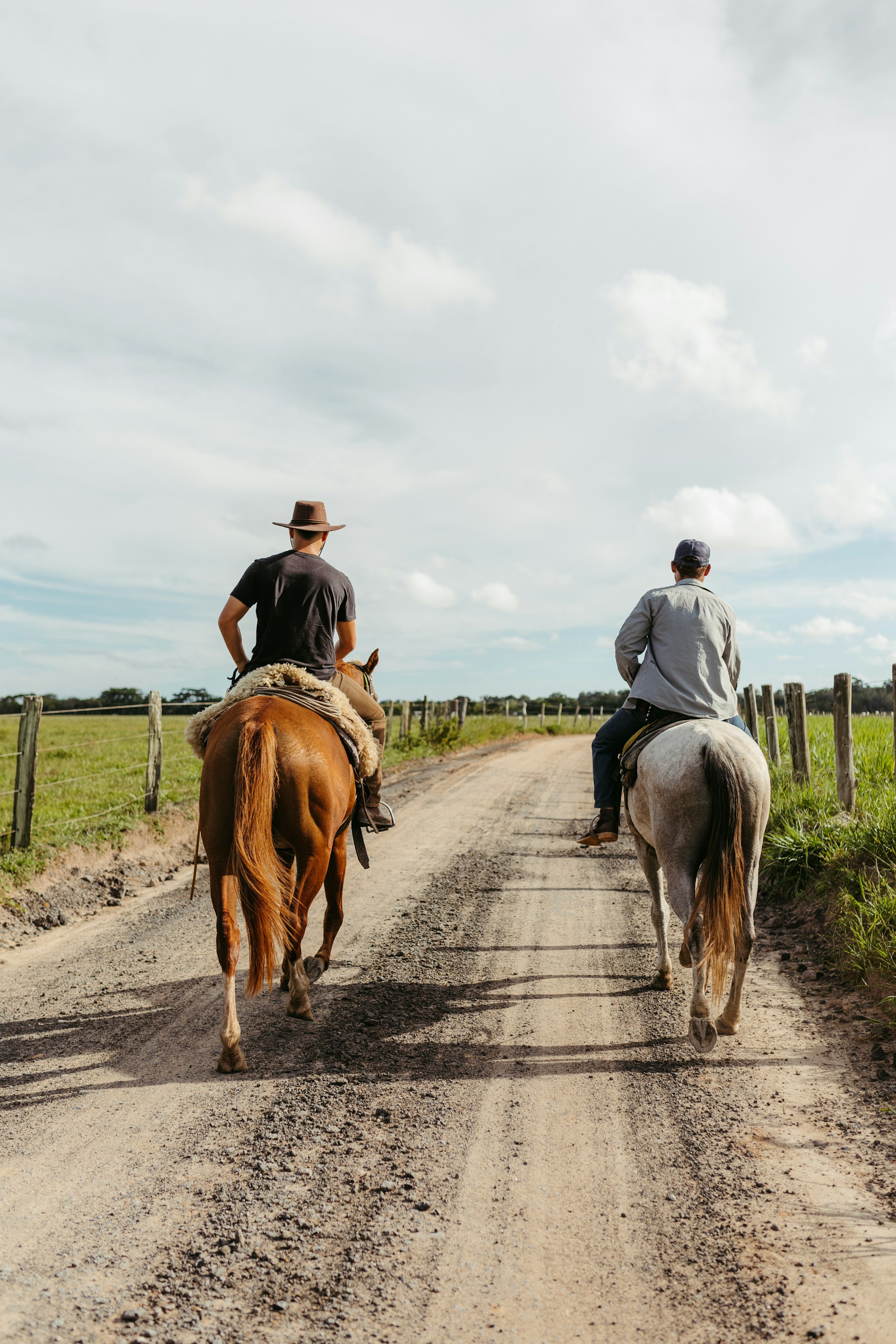 Two people riding horses on a rural dirt road surrounded by green pastures under a cloudy sky.