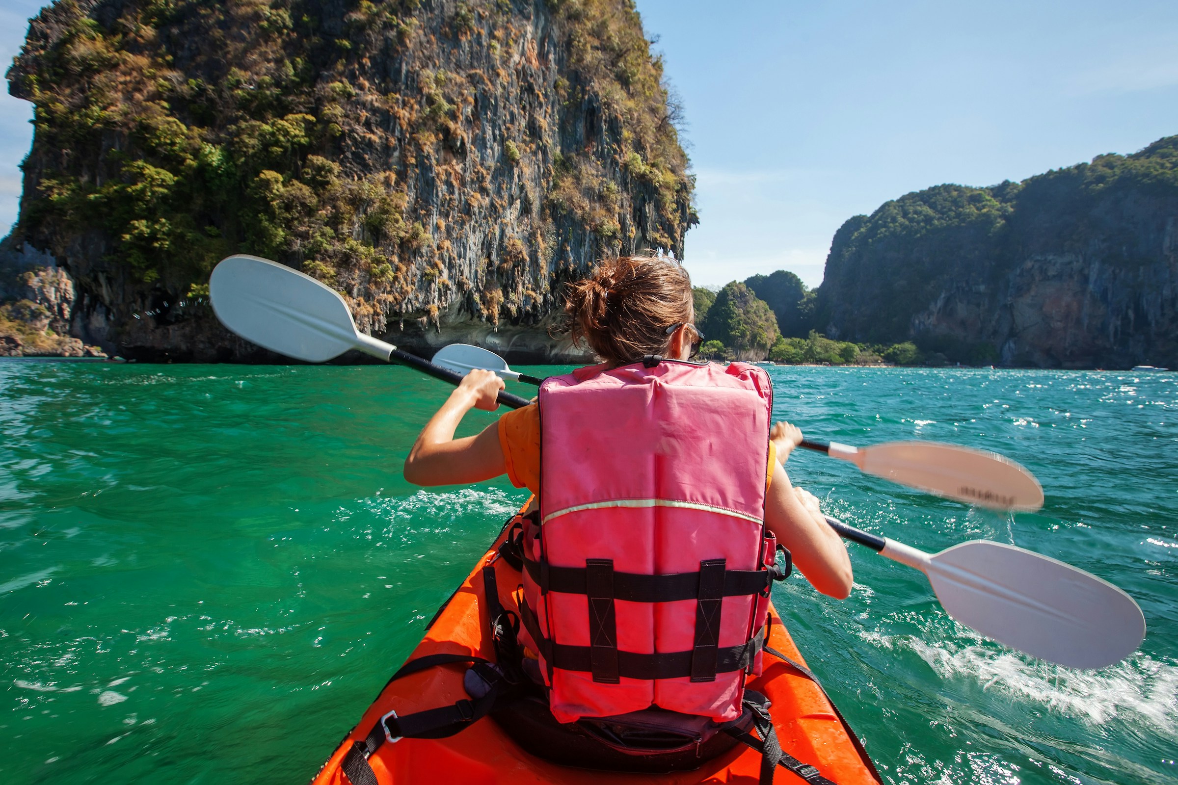 Person kayaking on turquoise water near rocky cliffs, wearing a pink life vest and paddling an orange kayak under clear blue skies.