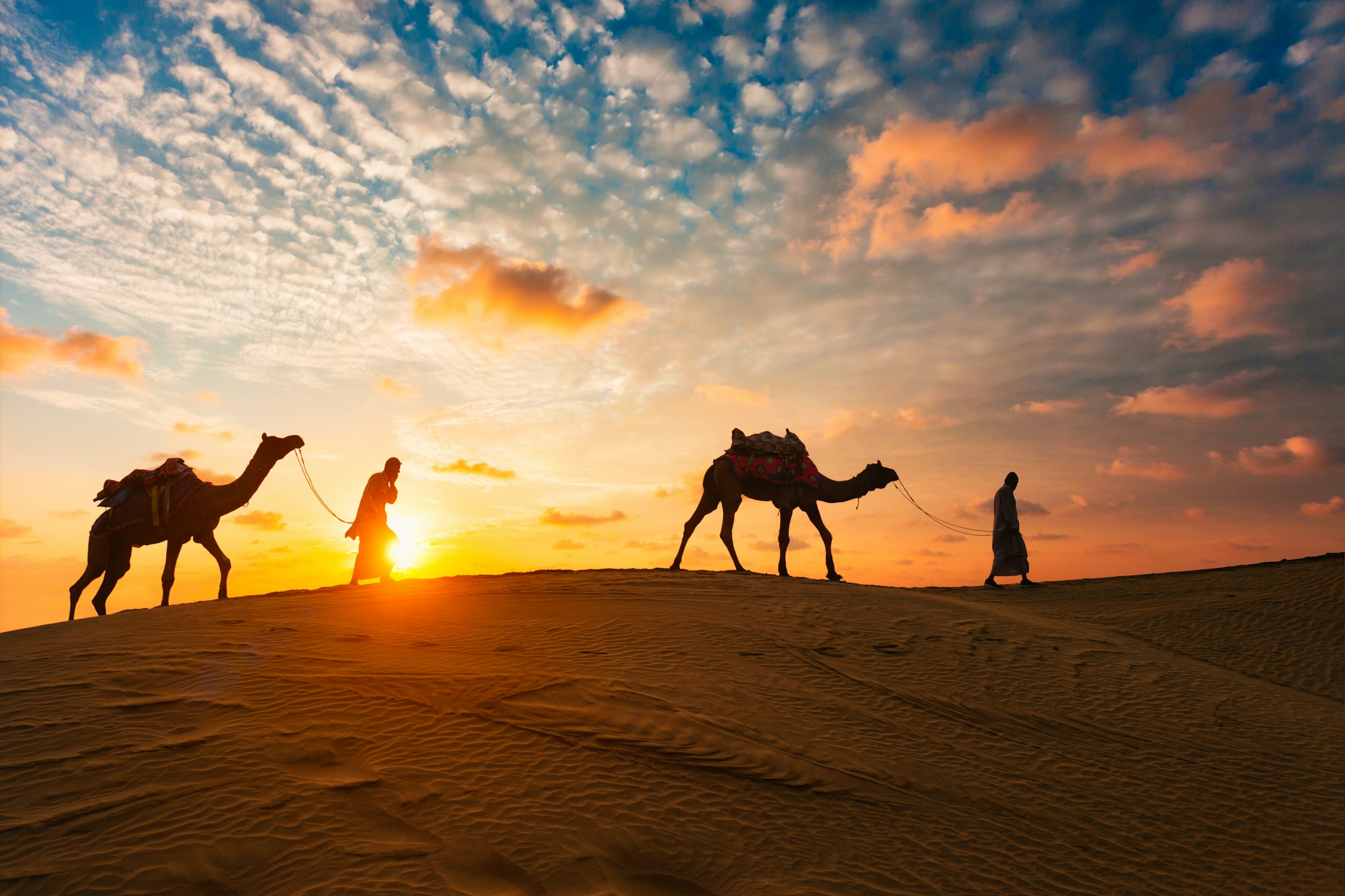 Silhouettes of two people leading camels across a desert at sunset, with a vibrant sky filled with colorful clouds.