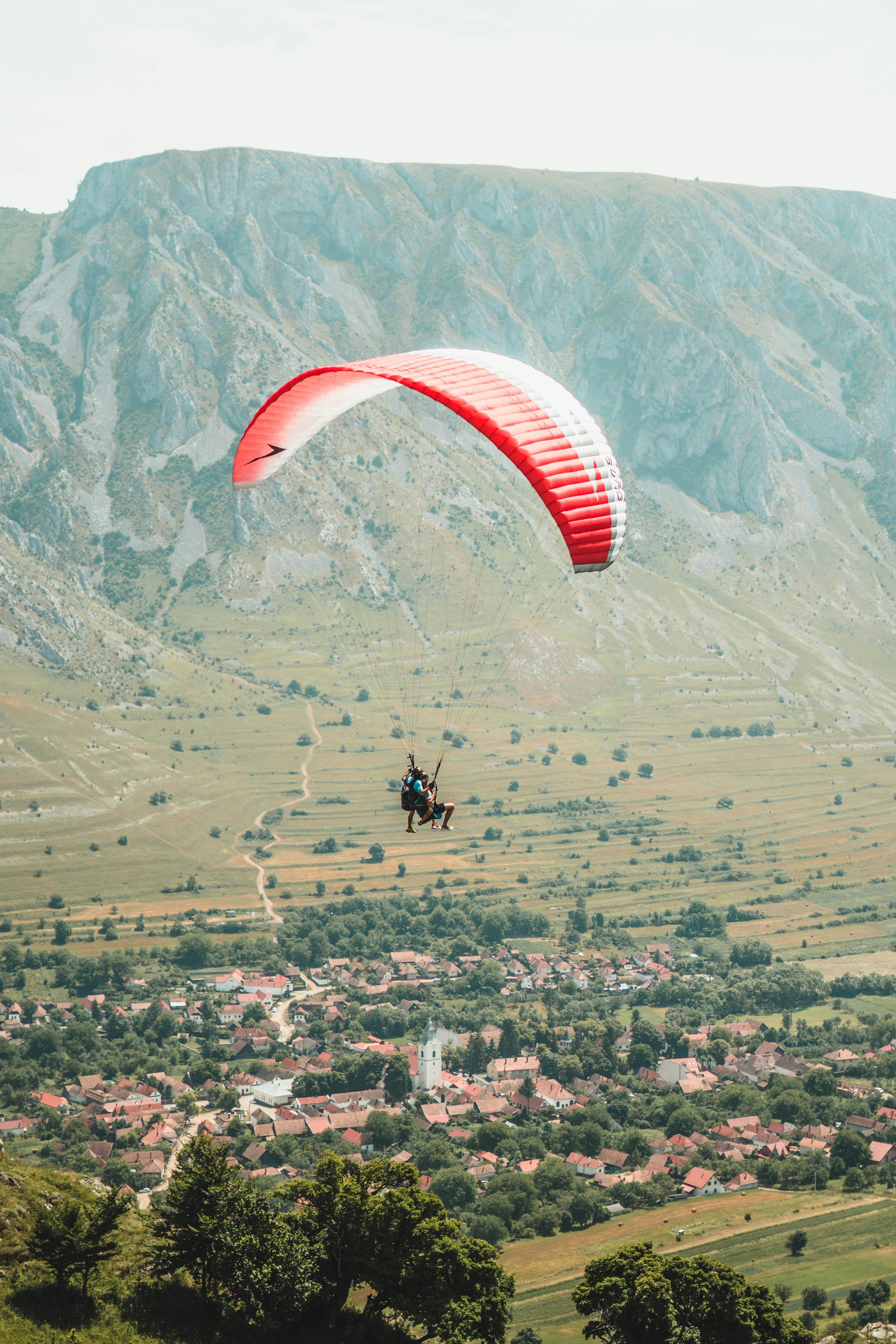 Paraglider flying over a scenic rural landscape with a small village and rolling hills below.