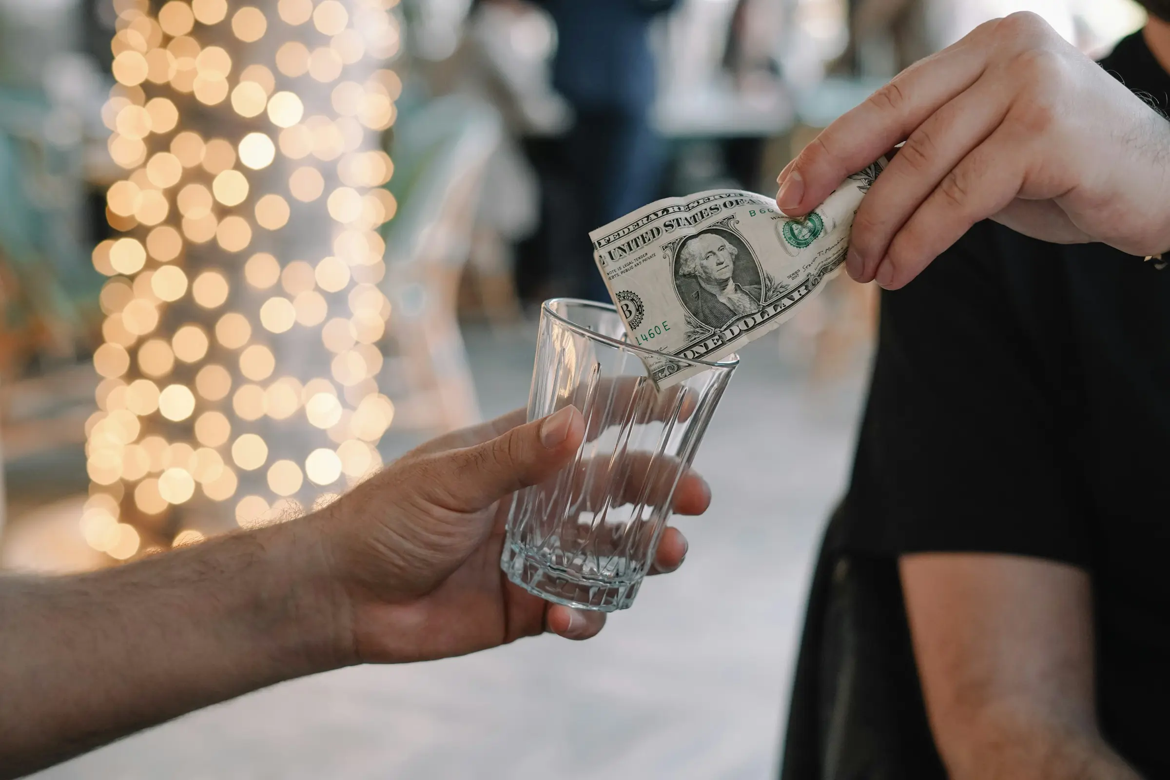 Person placing a one dollar bill into an empty glass at an event with string lights in the background.
