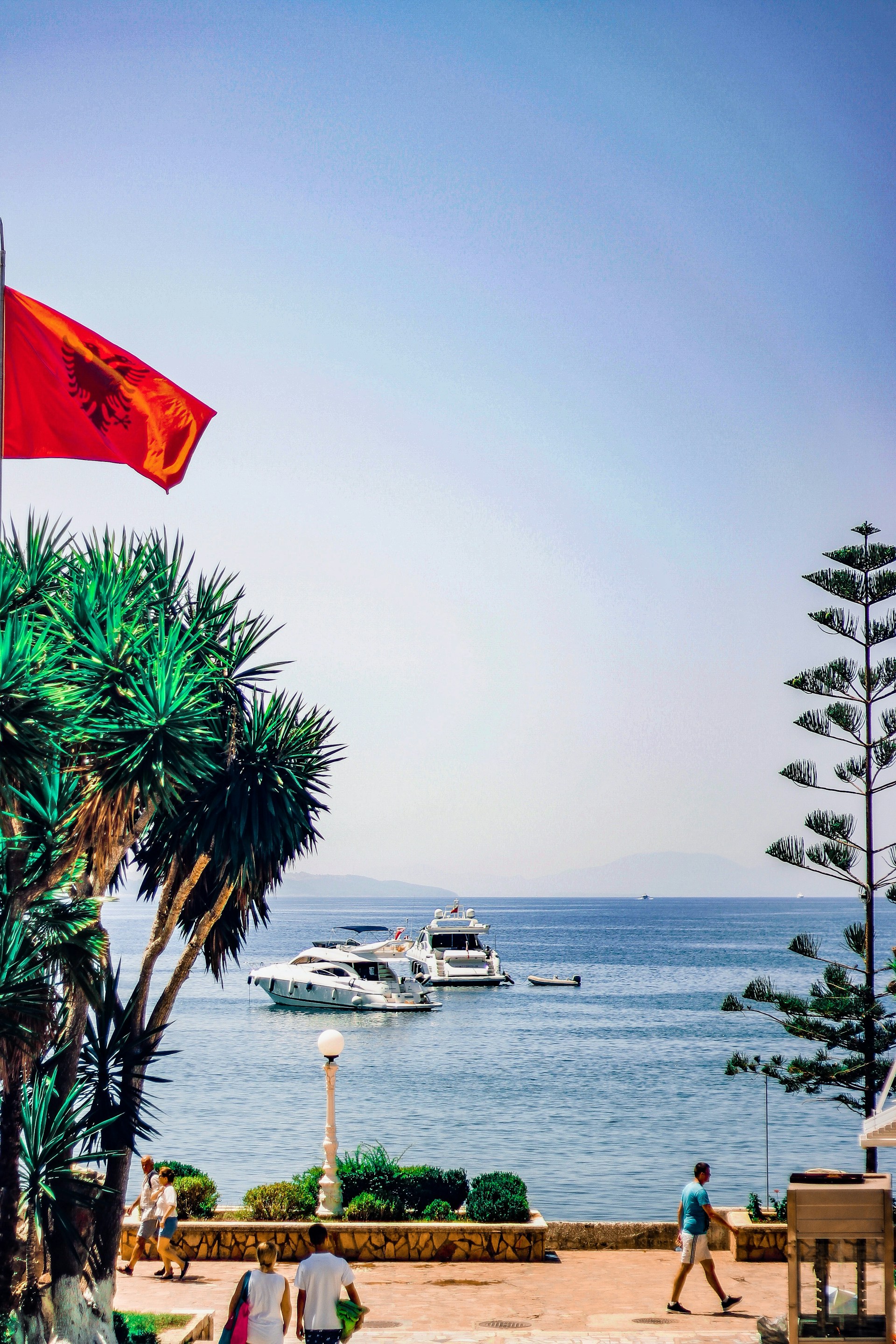 Röd flagga vajar över en strandpromenad vid havet med båtar och palmer i förgrunden, soligt väder