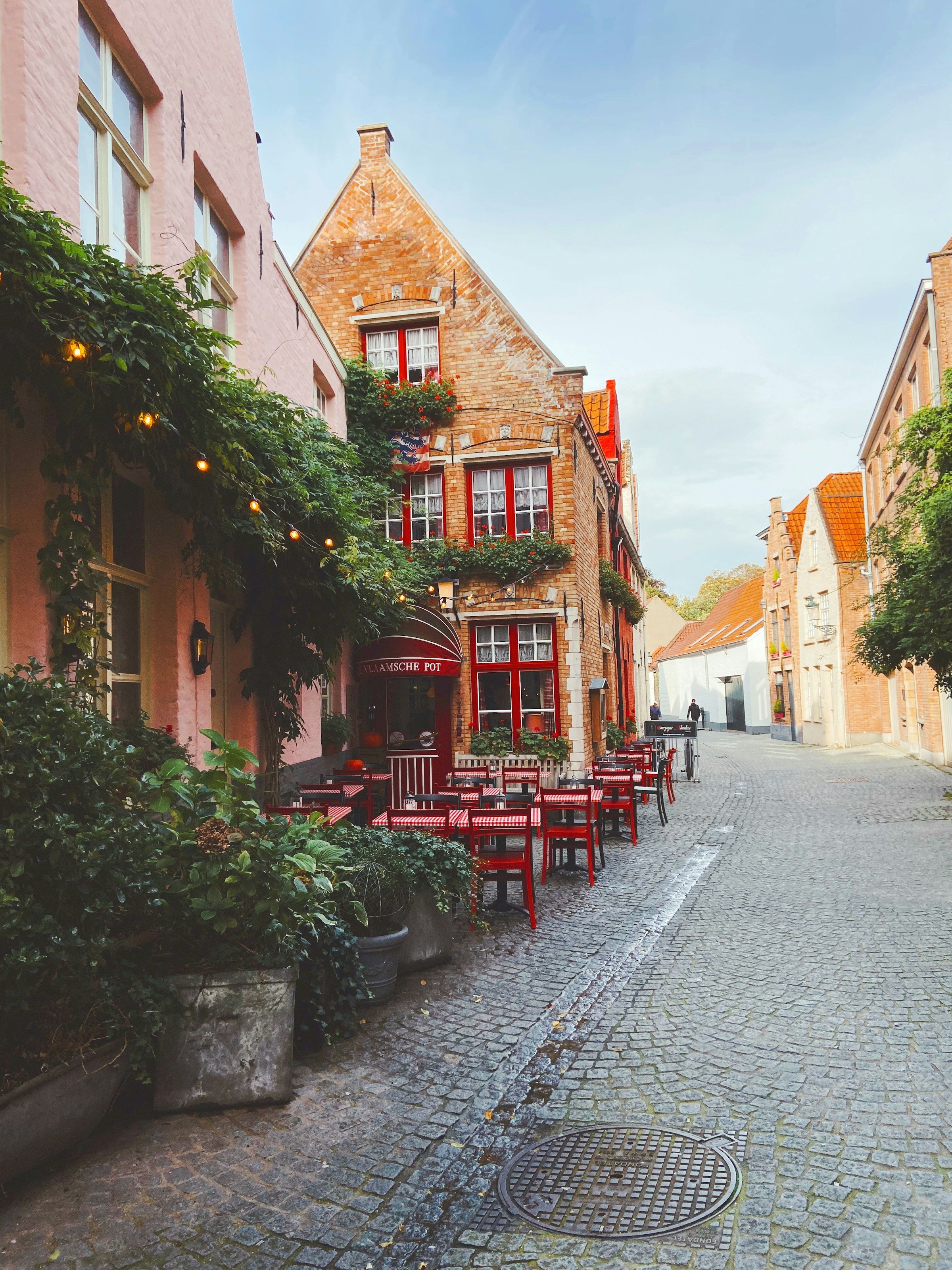 Charming European cobblestone street with historic brick buildings, ivy-covered walls, and a quaint outdoor cafe with red chairs and tables.