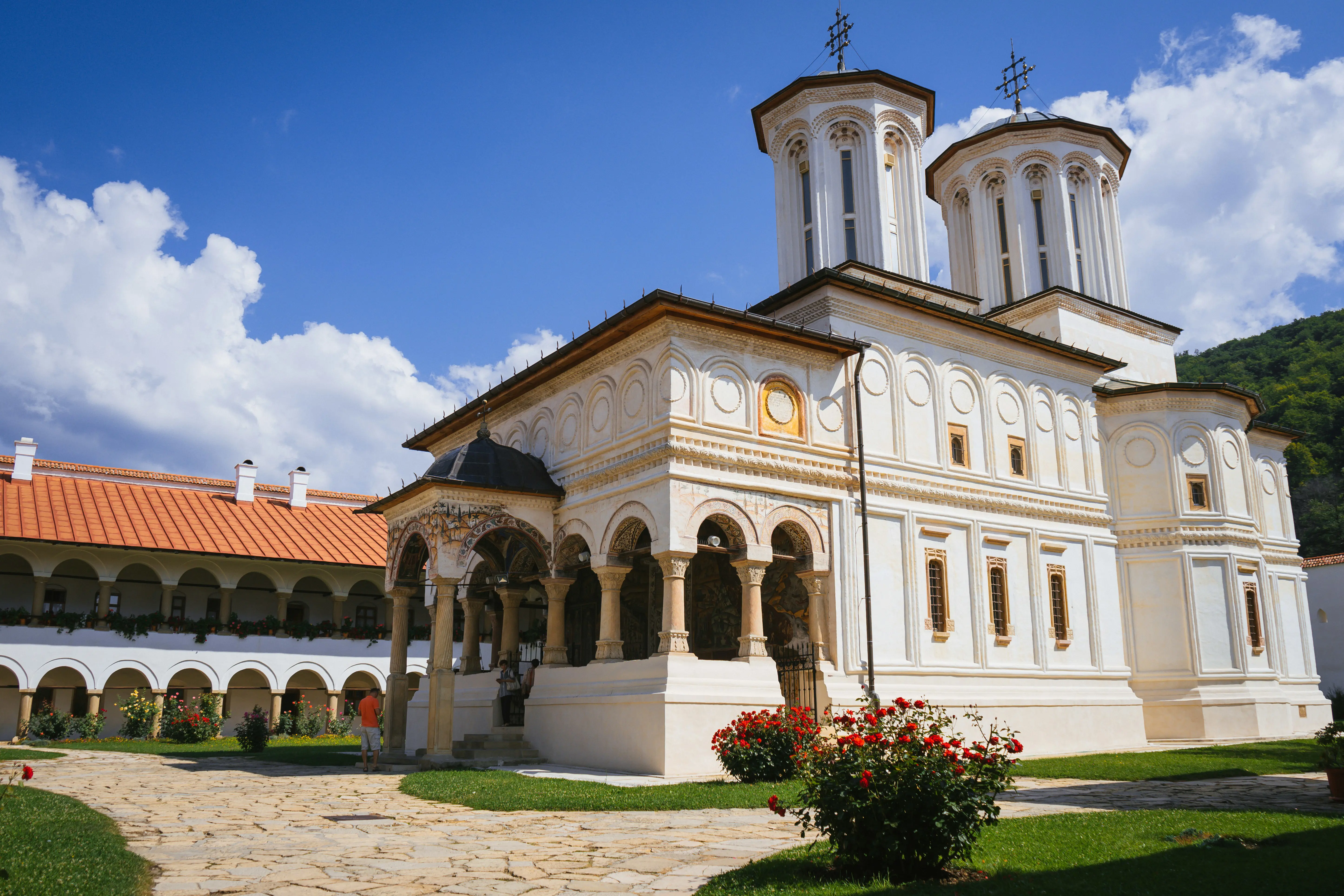 Historic white church with ornate carvings and tall towers under a blue sky, surrounded by a courtyard with flowers and arches, located in a scenic, green landscape.