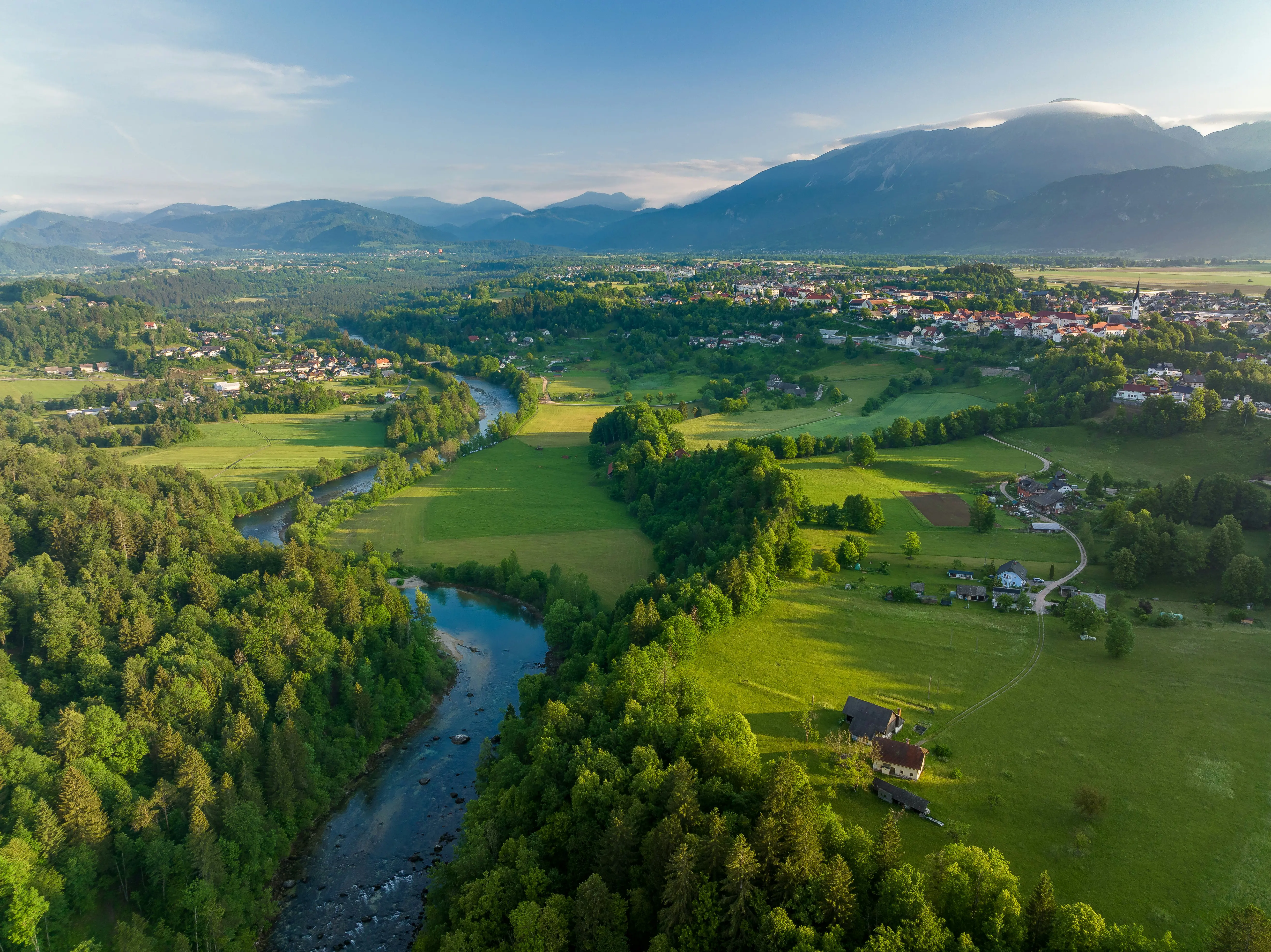Aerial view of a lush green valley with a winding river and distant mountains, featuring a small village surrounded by forests in Bosnia.
