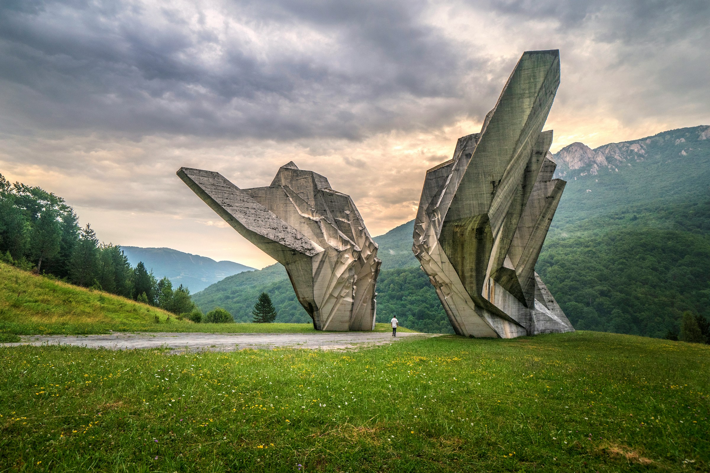 Brutalist stone monument in the Valley of Heroes at Tjentište, surrounded by lush green hills and overcast sky.
