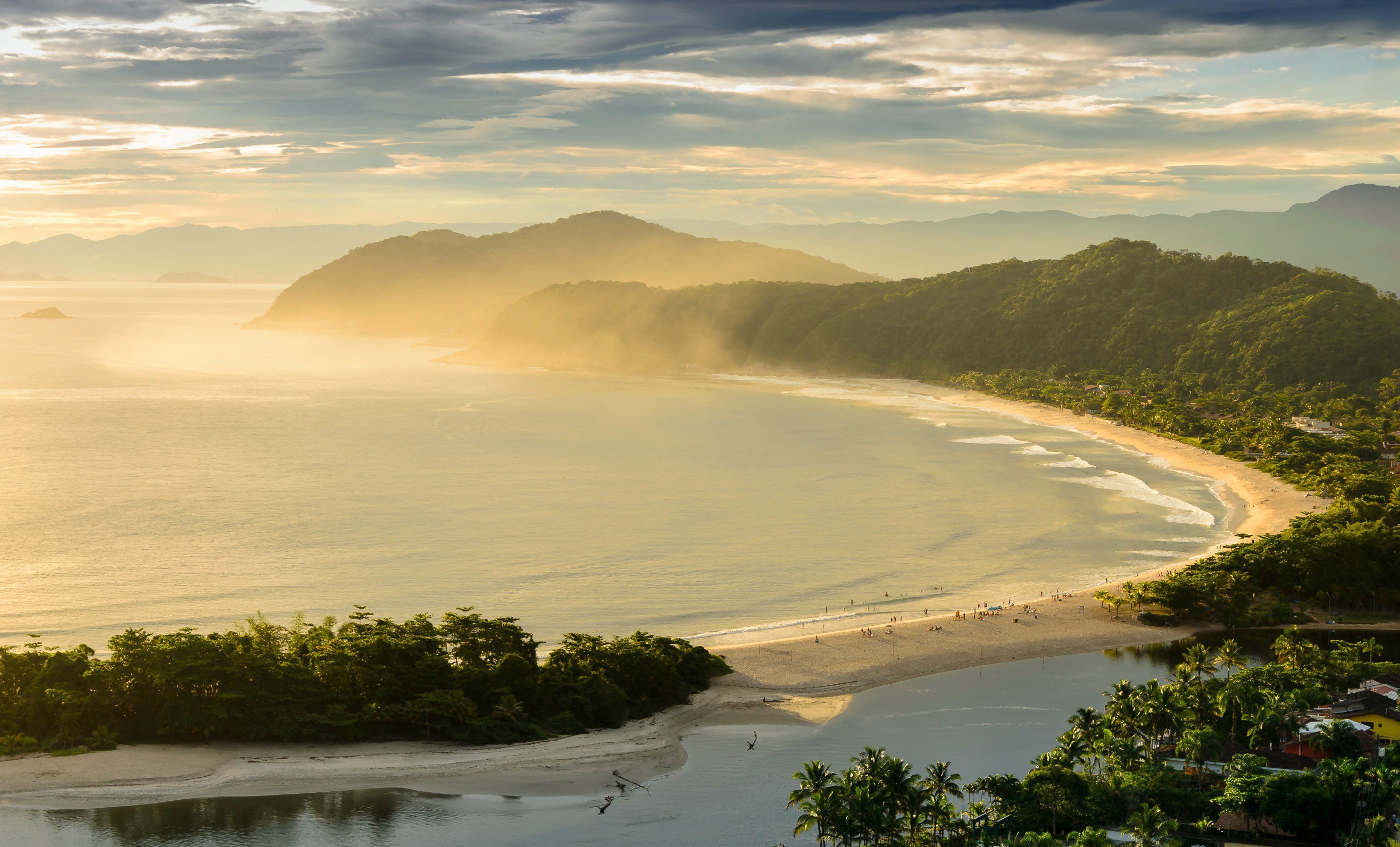 Travel to Brazil - Serene tropical beach with lush green hills and a golden sunset sky, featuring calm ocean waves and a sandy shoreline lined with palm trees.