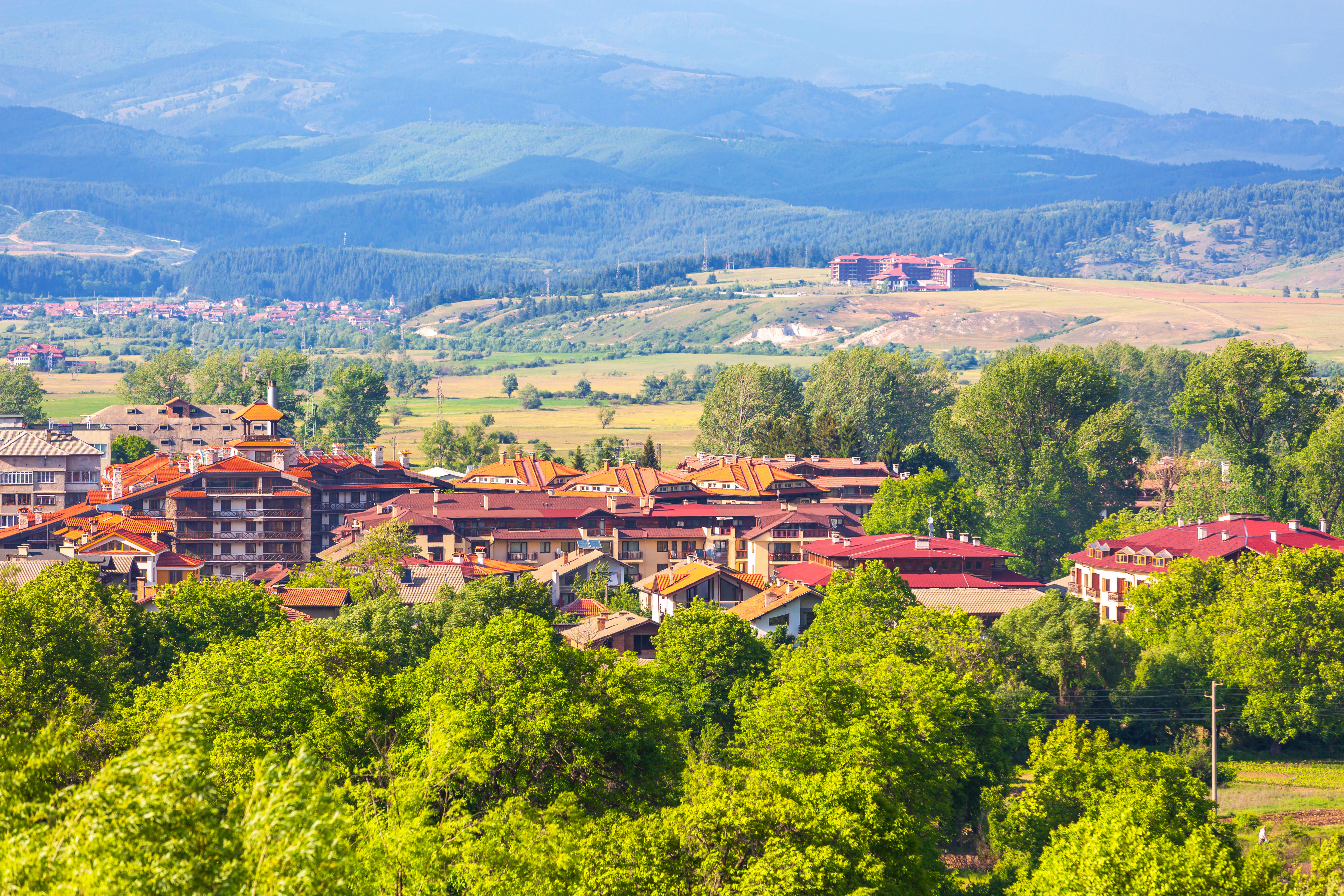 Kesäinen maisema vuoristoisesta kylästä, jossa on punakattoisia rakennuksia ja vihreää puustoa etualalla. Taustalla näkyy metsää ja mäkiä