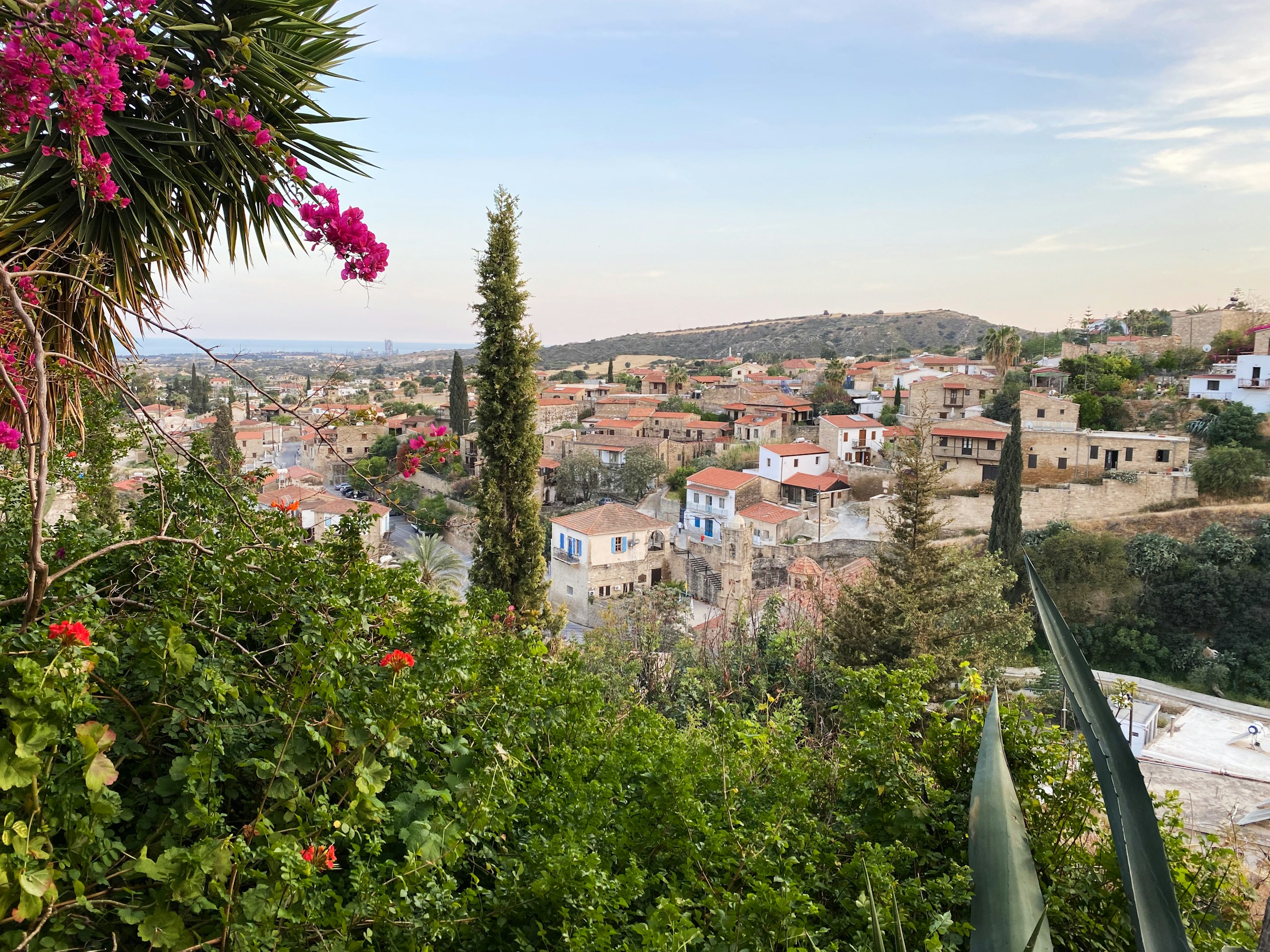 View of a traditional village in Larnaca with stone houses and red-tiled roofs, surrounded by lush greenery and colorful flowers.