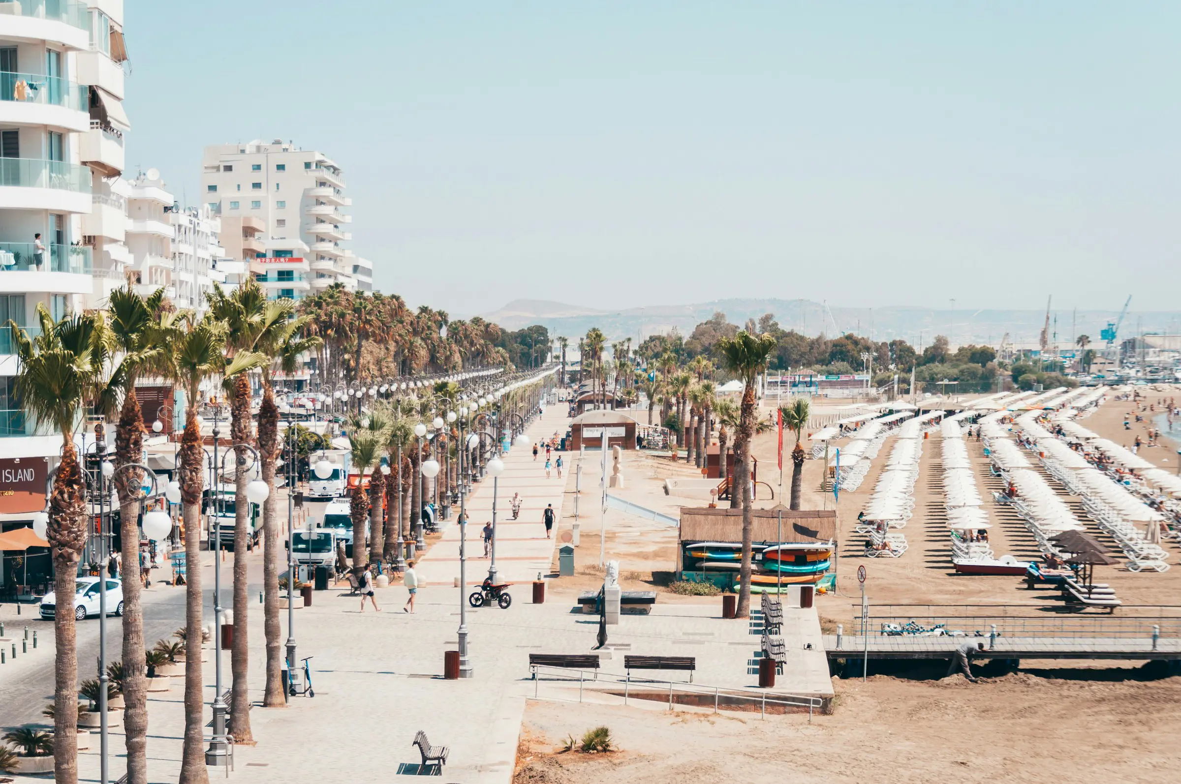 Beach promenade with palm trees and high-rise buildings in Larnaca, Cyprus, featuring sunbathers under umbrellas along a sandy beach.