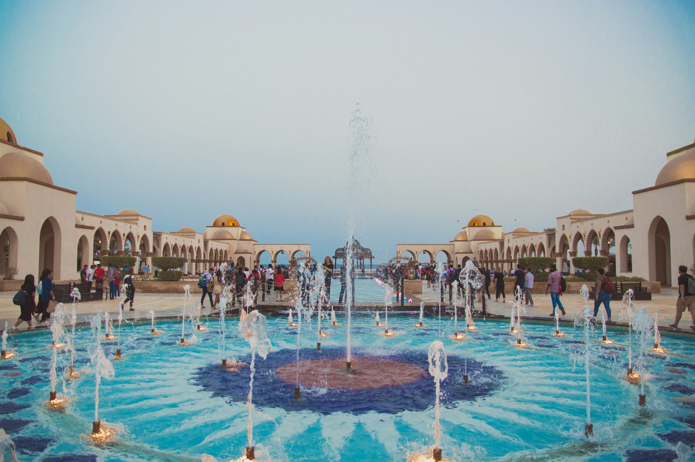 Majestic courtyard with vibrant fountain surrounded by domed architecture and crowds enjoying the scenery.