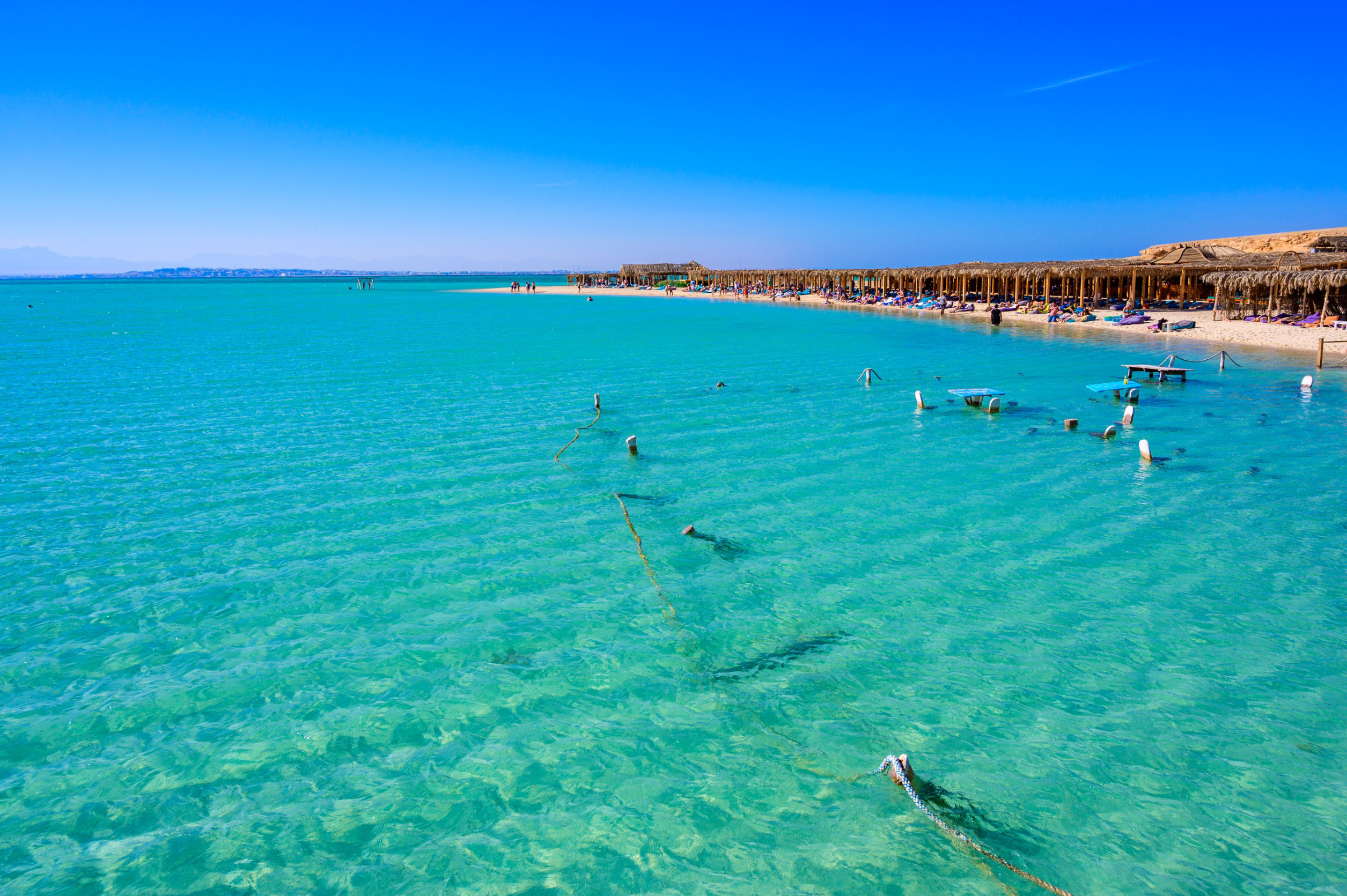 Travel to Hurghada - Beautiful tropical beach with clear turquoise water, sandy shore, and thatched beach umbrellas on a sunny day.