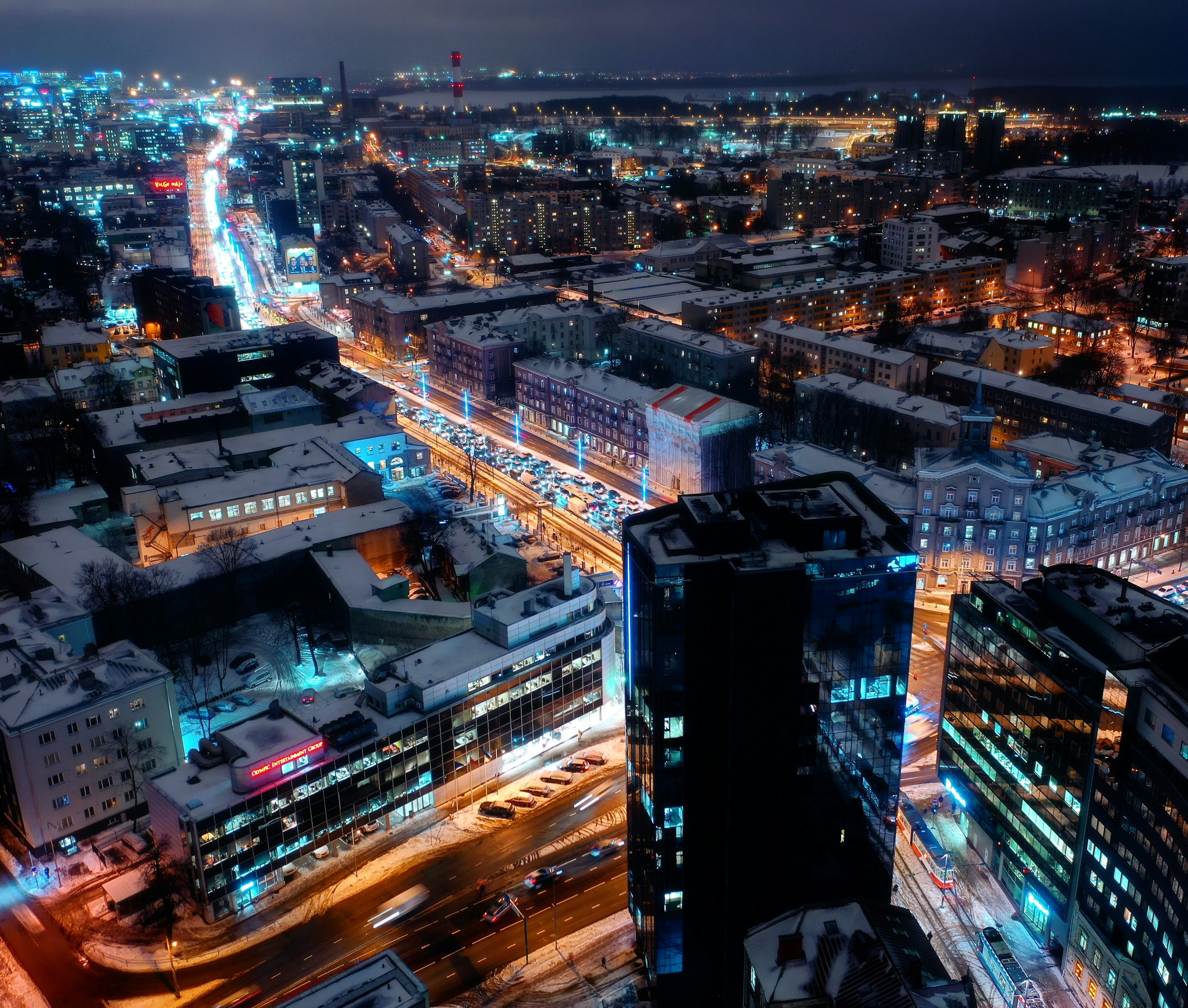 Aerial view of a vibrant cityscape at night, showcasing illuminated streets, modern high-rise buildings, and a bustling urban atmosphere, with snow-covered rooftops.