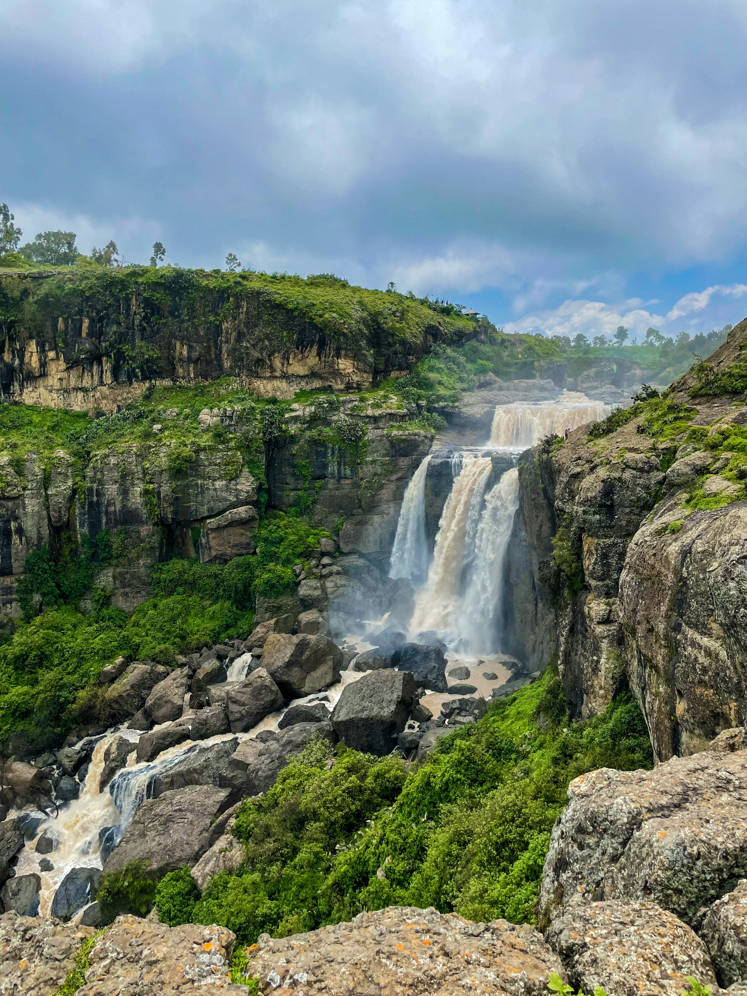 A scenic waterfall cascading over a cliff surrounded by lush greenery and rocky terrain under a cloudy sky.