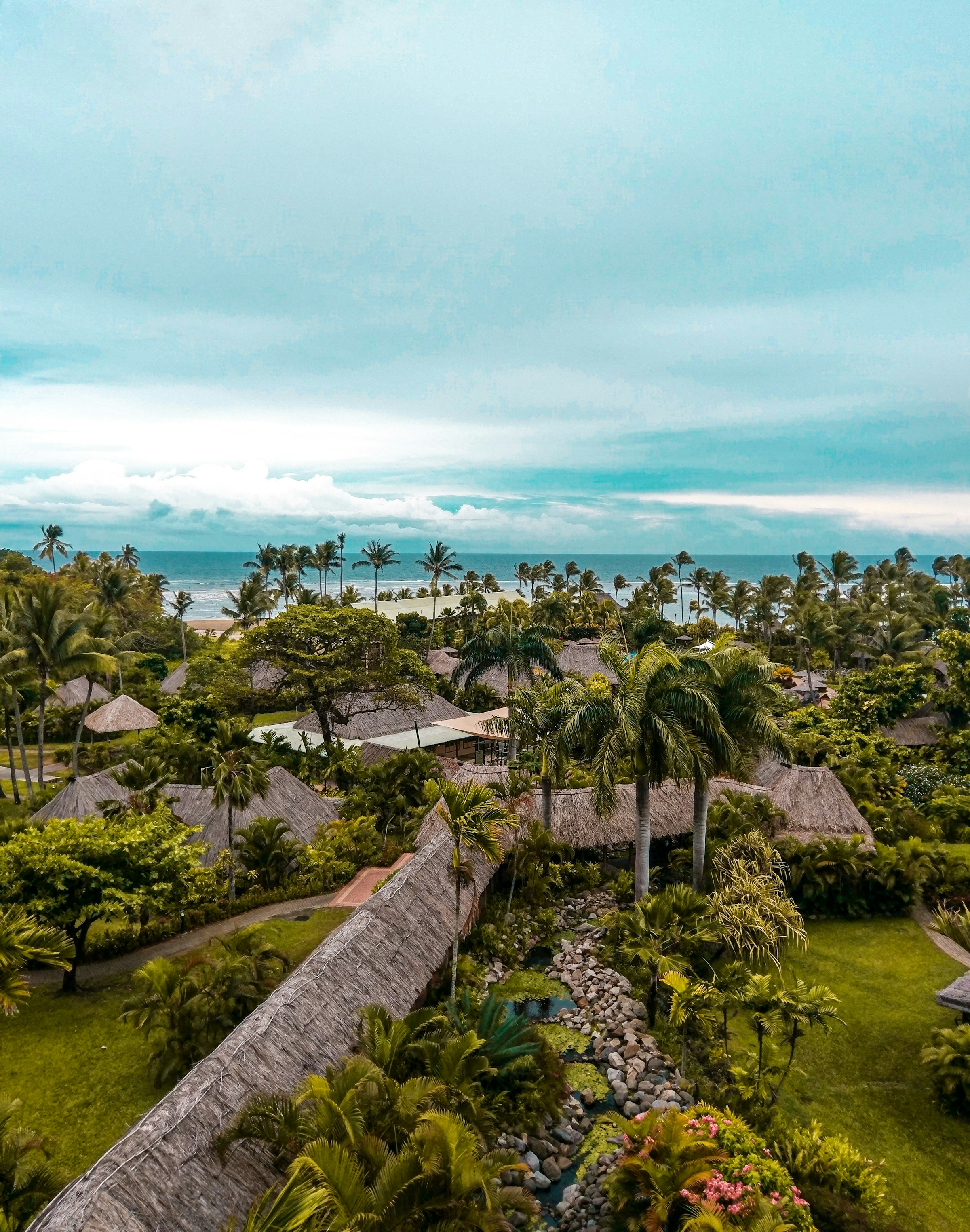Tropical resort with thatched-roof huts surrounded by lush greenery and palm trees, overlooking the ocean under a cloudy sky.