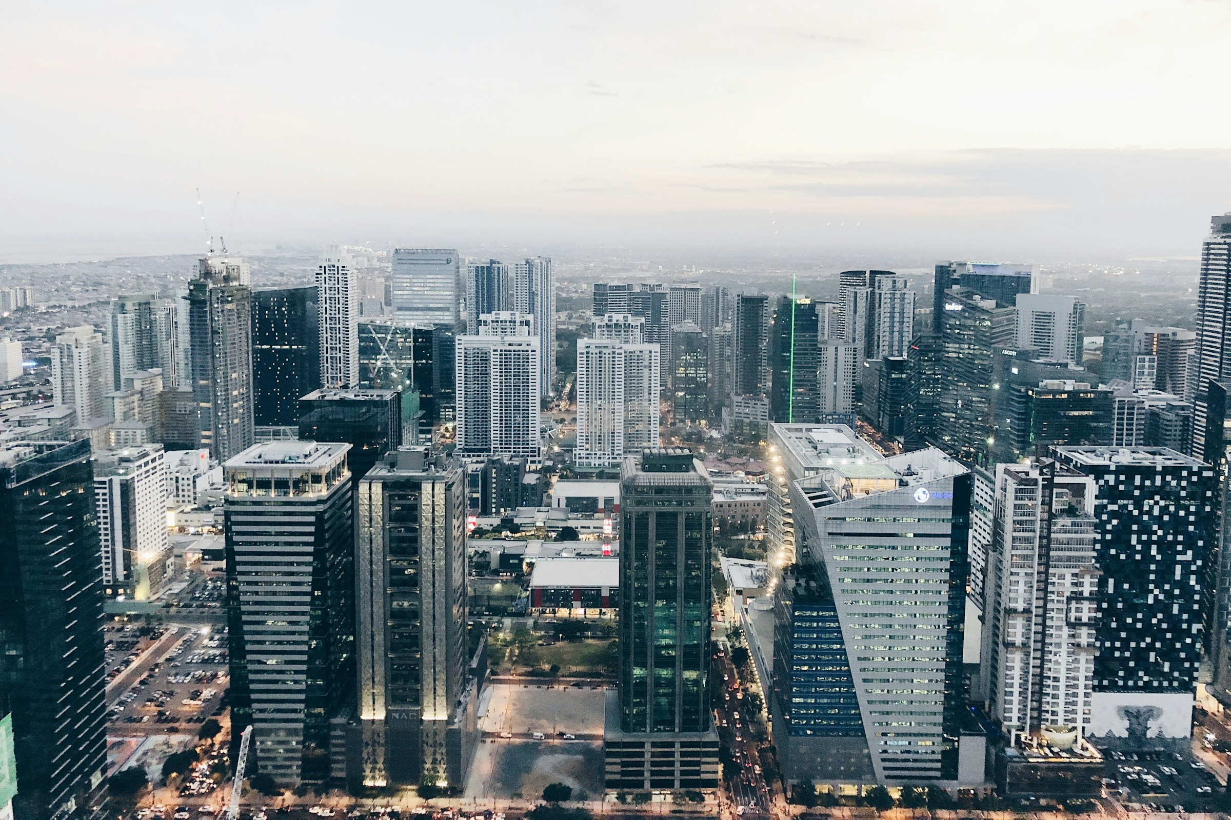 Skyline of Manilla with numerous skyscrapers at dusk, featuring illuminated buildings and a cloudy sky in the background.