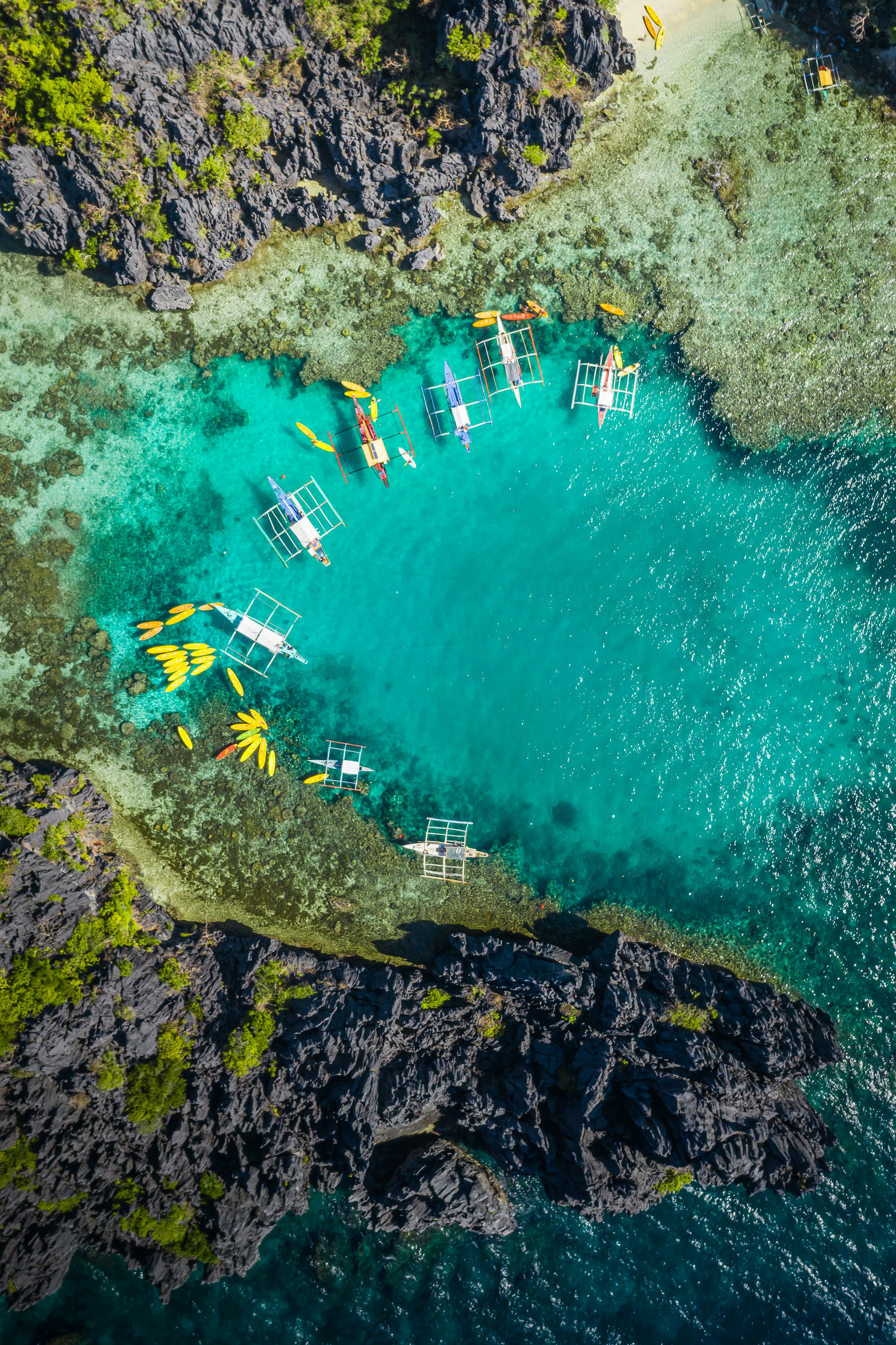 Aerial view of traditional outrigger boats in clear turquoise water near rocky cliffs and lush greenery in El Nido, Palawan, Philippines.
