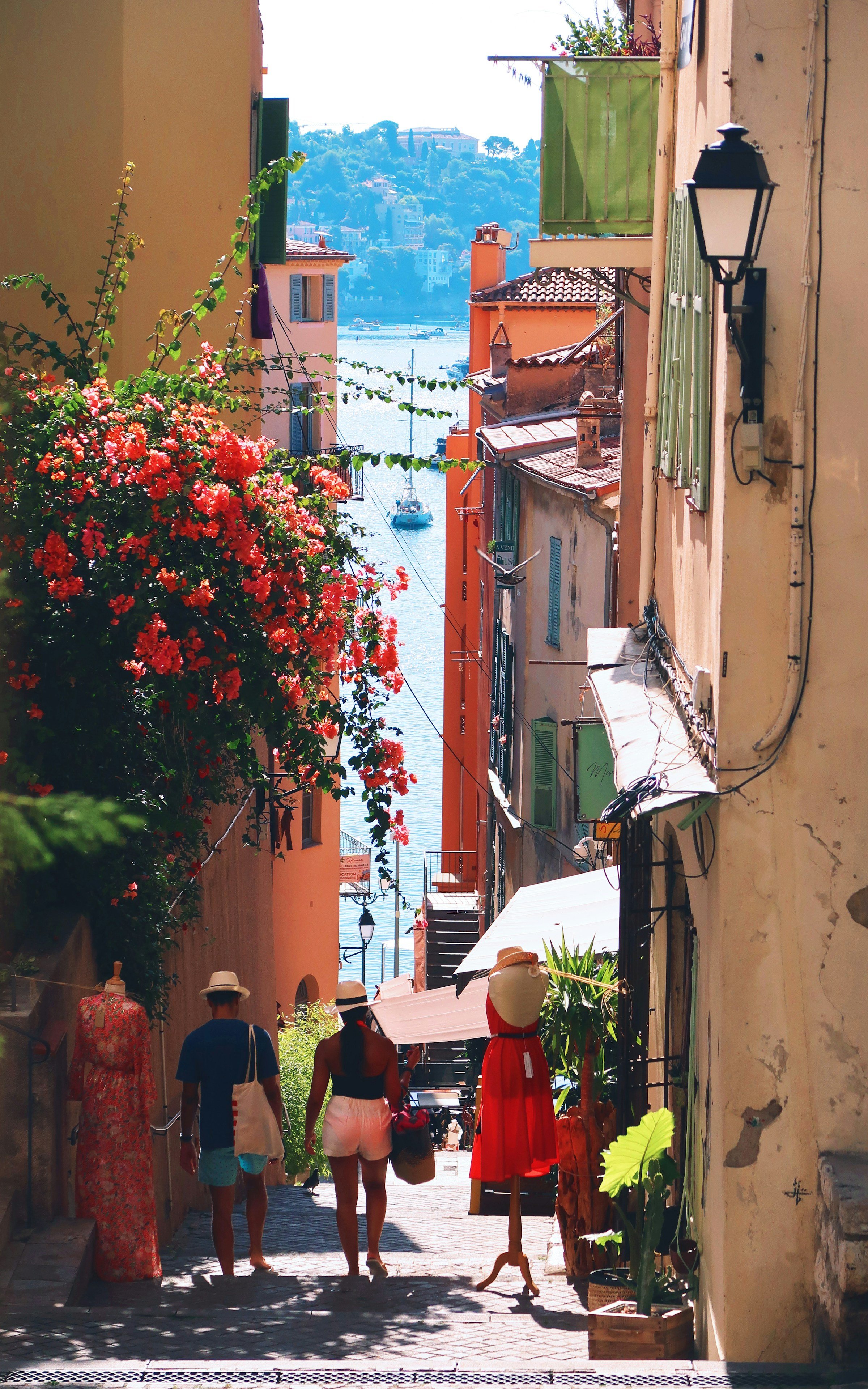 Narrow picturesque alley in a Mediterranean coastal village, with colorful buildings, flowering plants, and people strolling toward the sea.