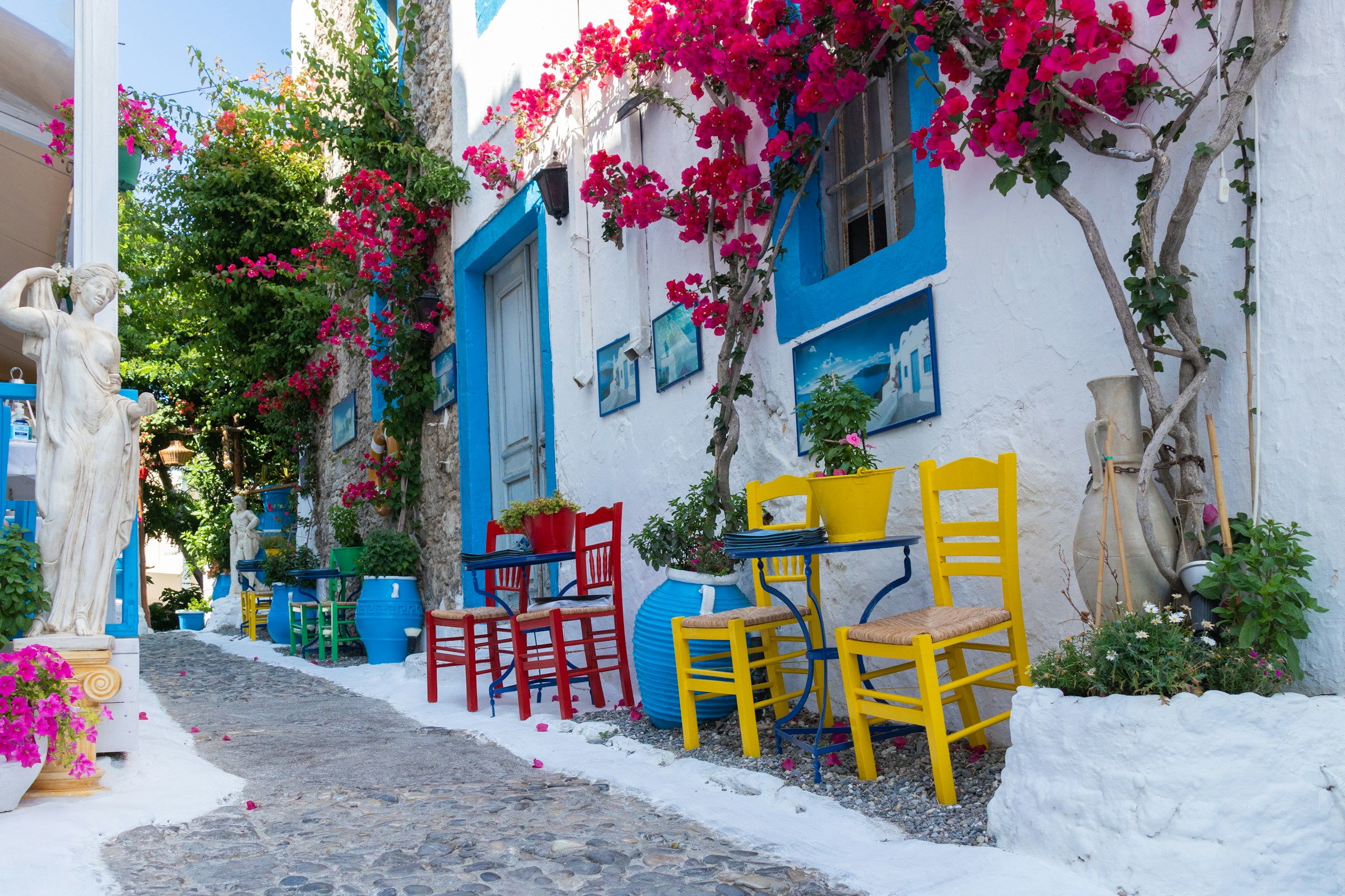 Colorful Greek street with vibrant flowers, blue-framed windows, red and yellow chairs, and a white statue amidst traditional architecture.