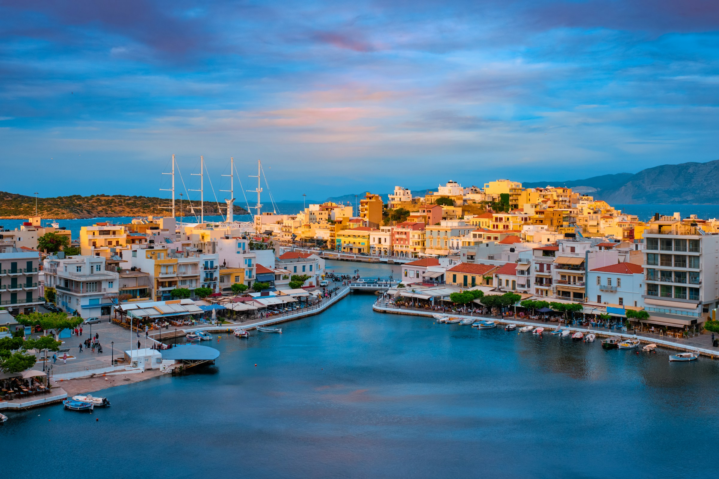 Scenic view of Agios Nikolaos, Crete, featuring colorful buildings along the waterfront, boats docked in the marina, and mountains in the background at sunset.