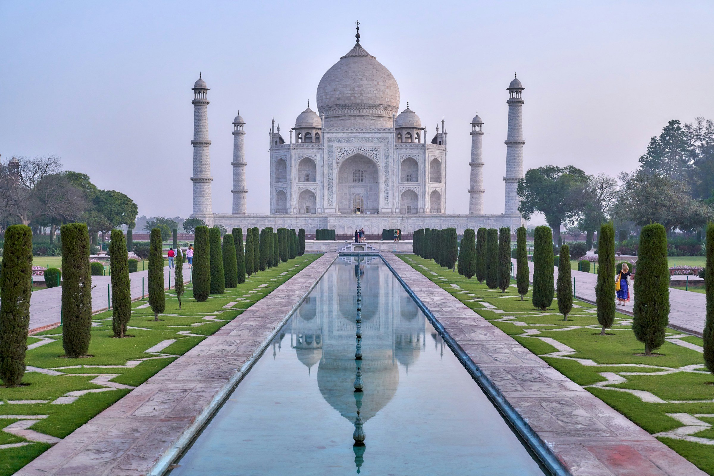 Taj Mahal at sunrise, reflecting in the central water channel, surrounded by lush gardens and tall trees in Agra, India.