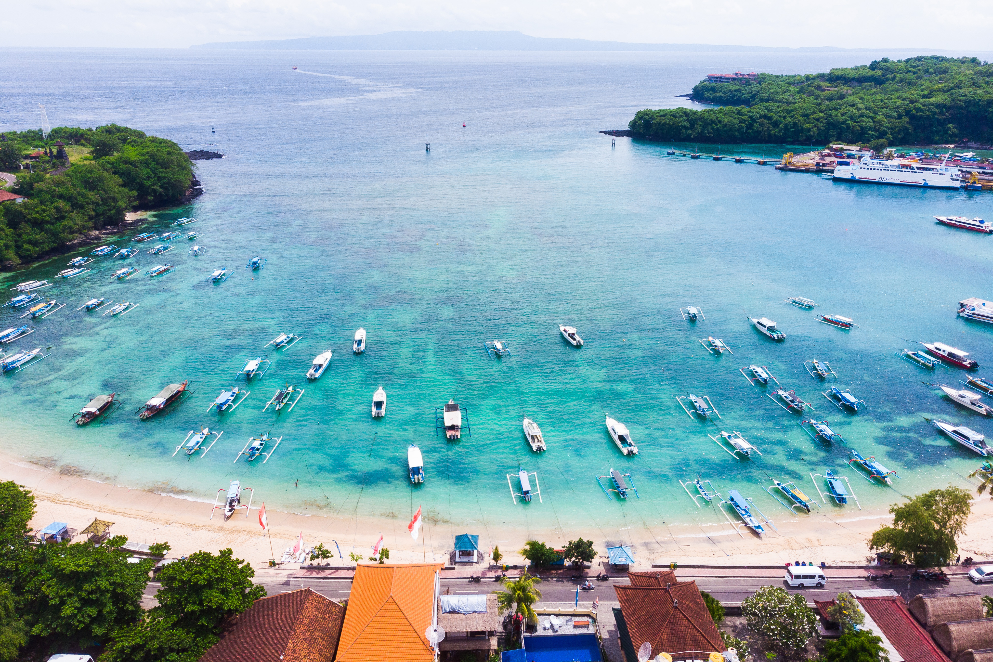 Vy över en tropisk strand med klart blått vatten och många båtar som ligger förtöjda vid kusten på Bali, omgiven av grönskande träd och byggnader