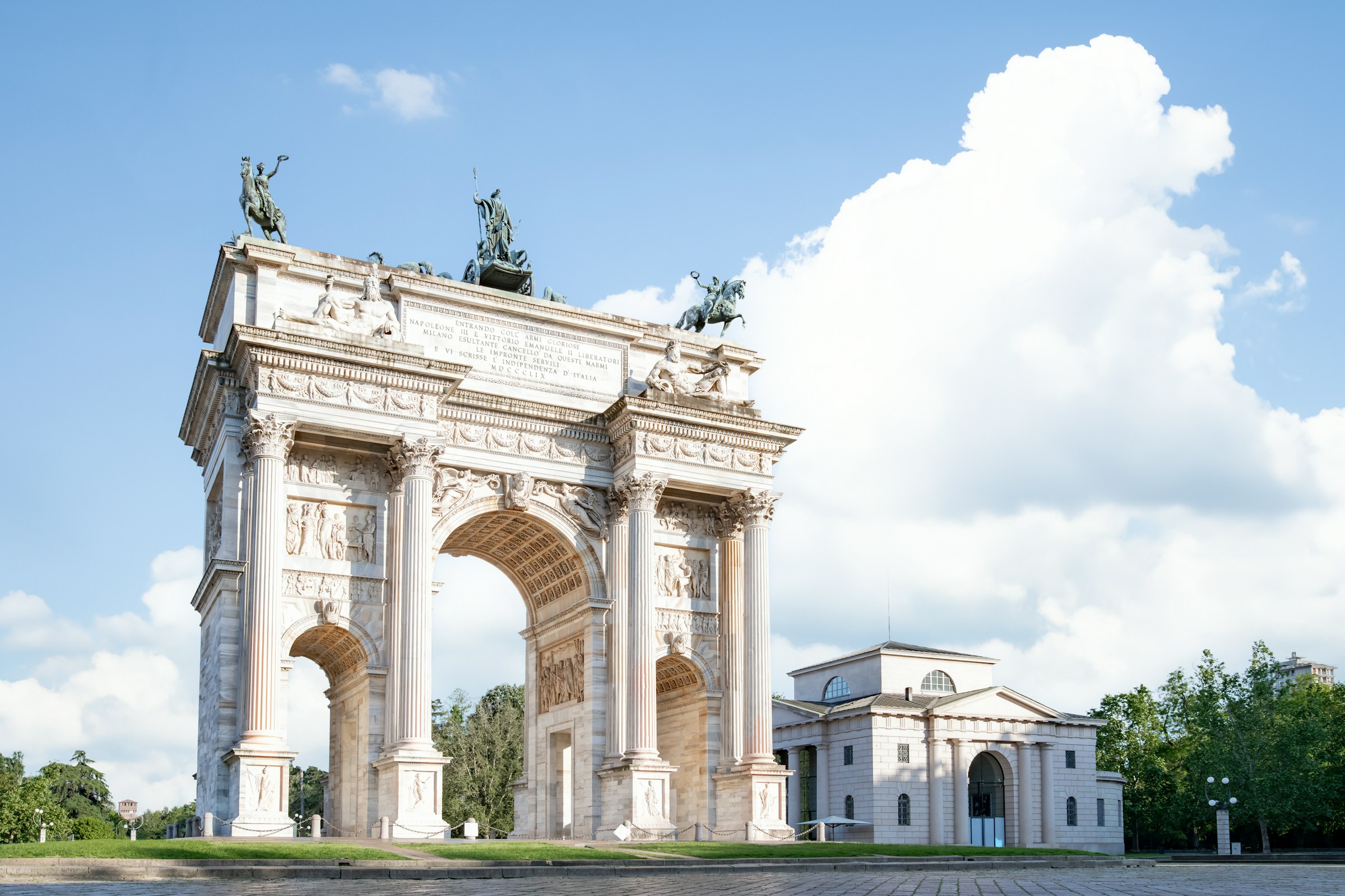 Triumphal Arch of Peace in Milan, Italy, under a clear blue sky with fluffy clouds, surrounded by greenery.