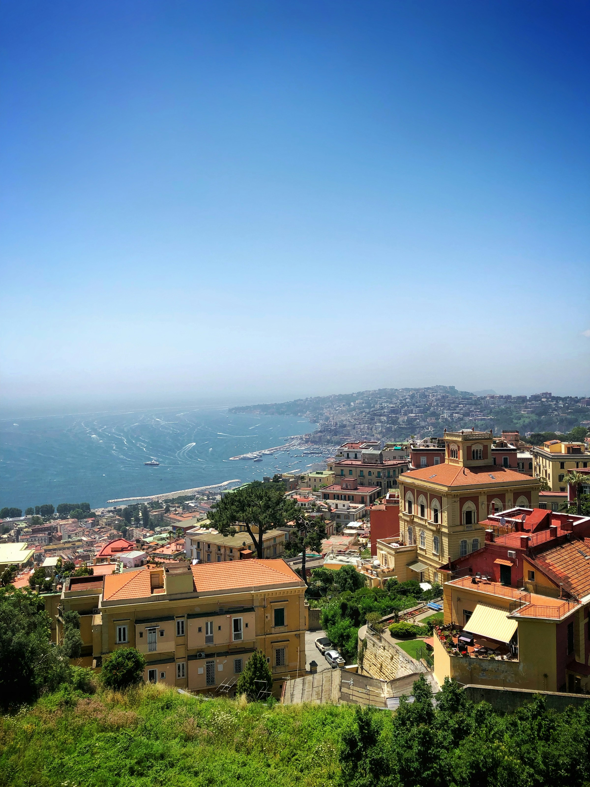 Scenic view of coastal cityscape in Naples, Italy, showcasing colorful buildings against a backdrop of a vibrant blue sea and clear sky.