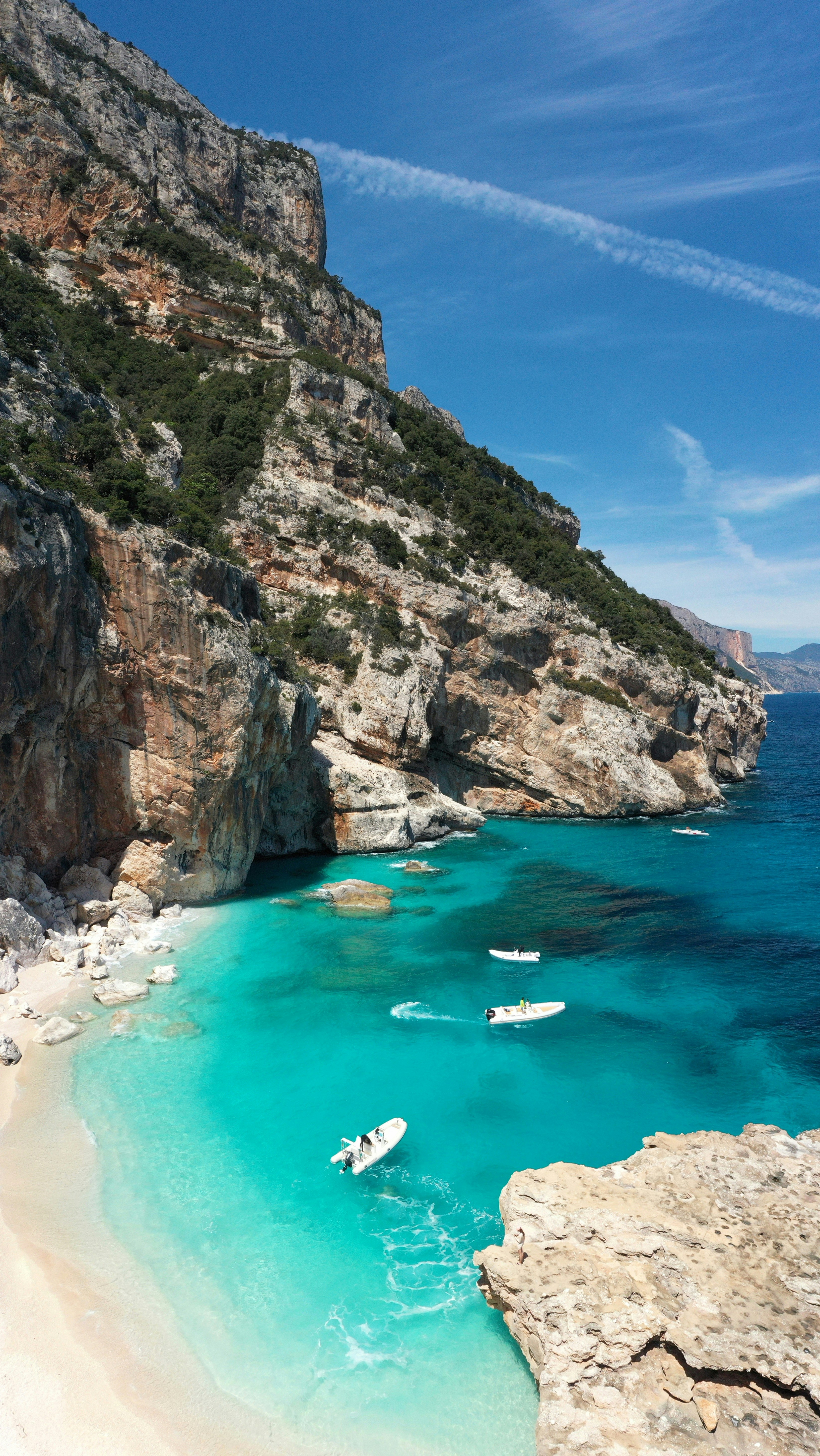 Klippor och turkost vatten vid en avskild strand med båtar vid Cala Mariolu, Sardinien, under en klarblå himmel