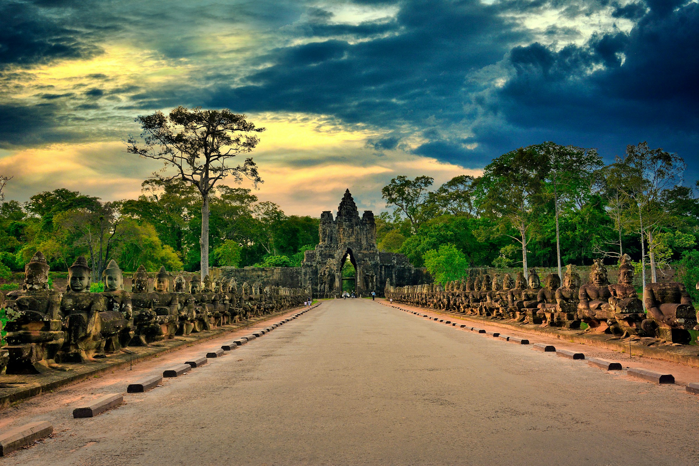 Ancient stone sculptures line the entrance to Angkor Thom in Cambodia, set against a dramatic sunset sky.