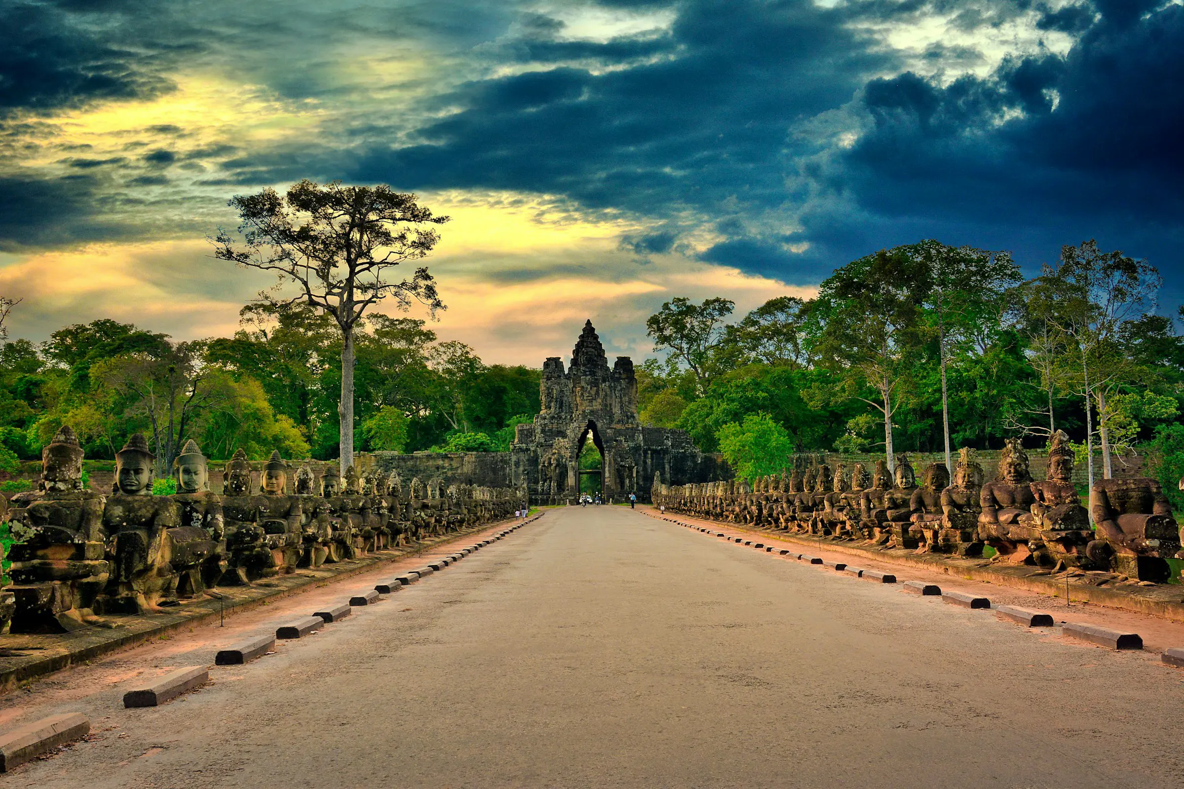 Ancient stone sculptures line the entrance to Angkor Thom in Cambodia, set against a dramatic sunset sky.