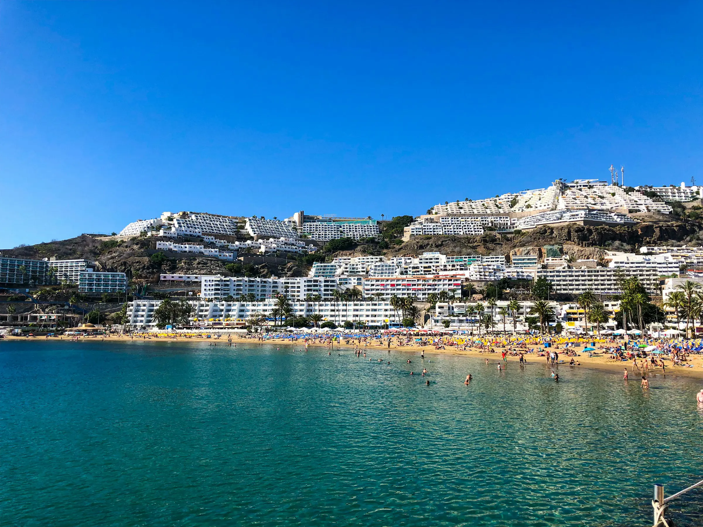 Strand och semesterort vid kusten på Gran Canaria, Kanarieöarna, Spanien, med turkost hav, sandstrand och vita höghus på en kulle under klarblå himmel.