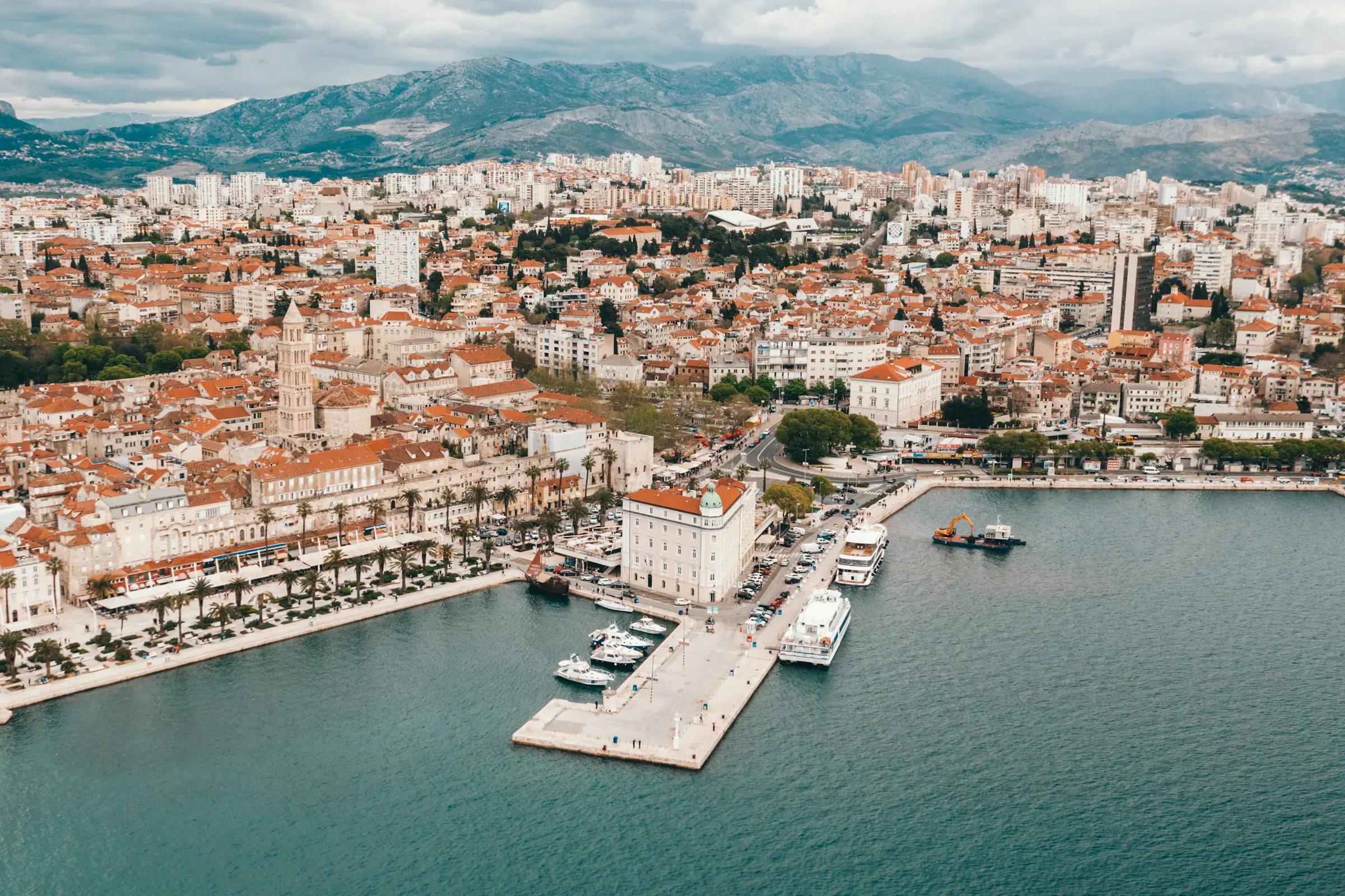 Aerial view of Split, Croatia, showcasing the historic cityscape with red-roofed buildings, waterfront promenade, and bustling harbor against a backdrop of mountains and cloudy skies