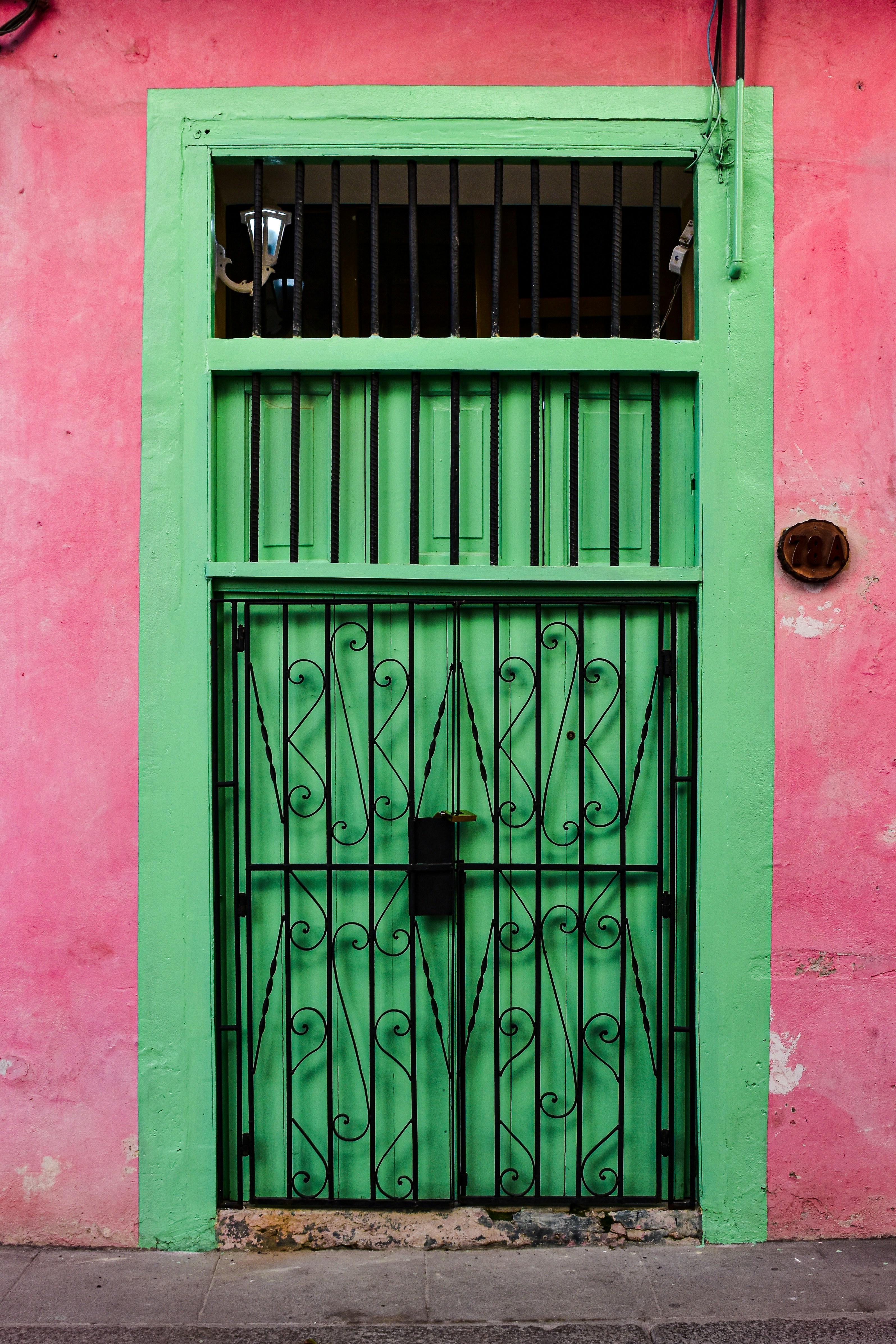 Eloisa vihreä metalliovi, jossa on koristeellinen musta rautaristikko, värikkään pinkkiä seinää vasten