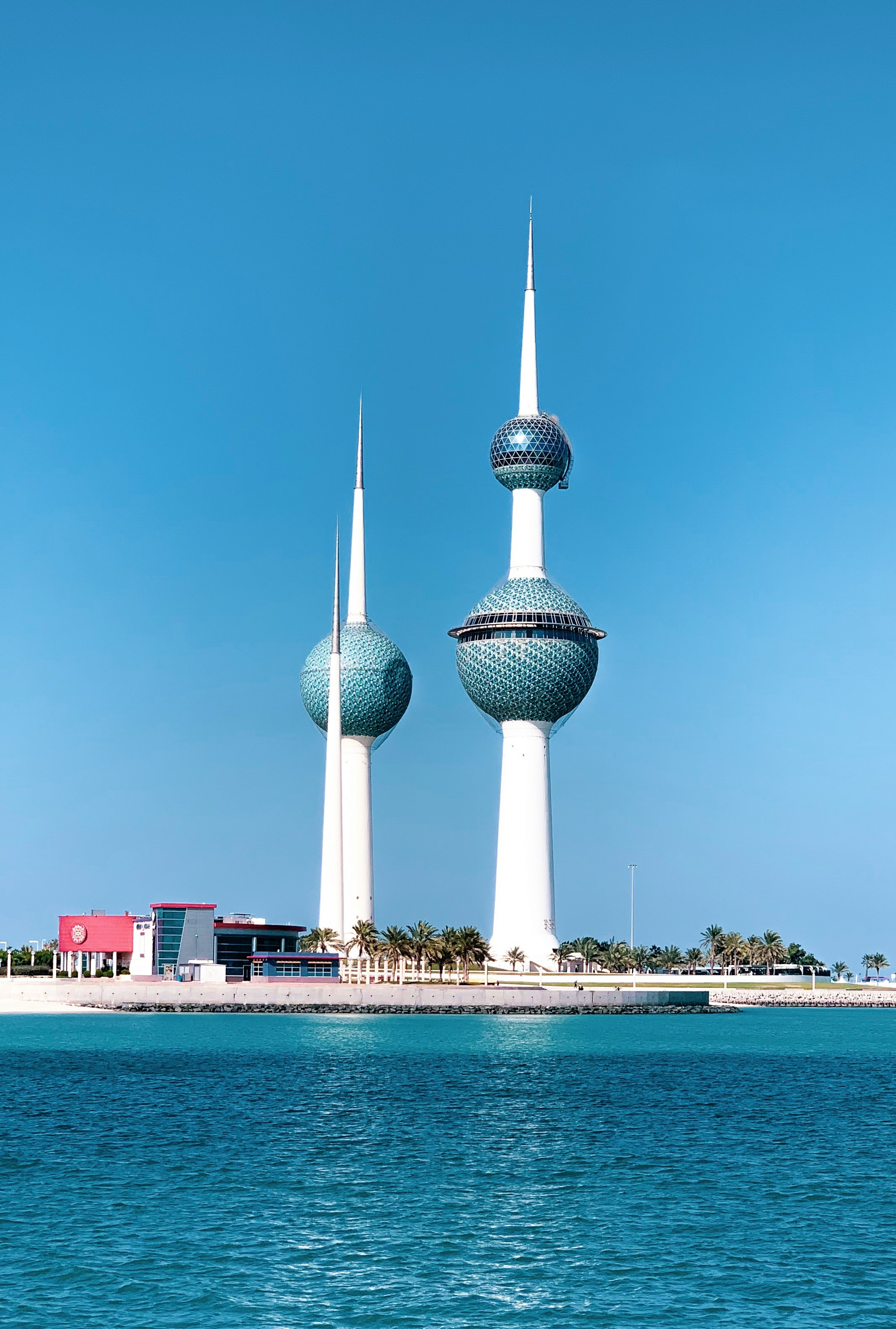 Kuwaittornen vid havet under en klarblå himmel, med moderna arkitektoniska strukturer och palmer i bakgrunden