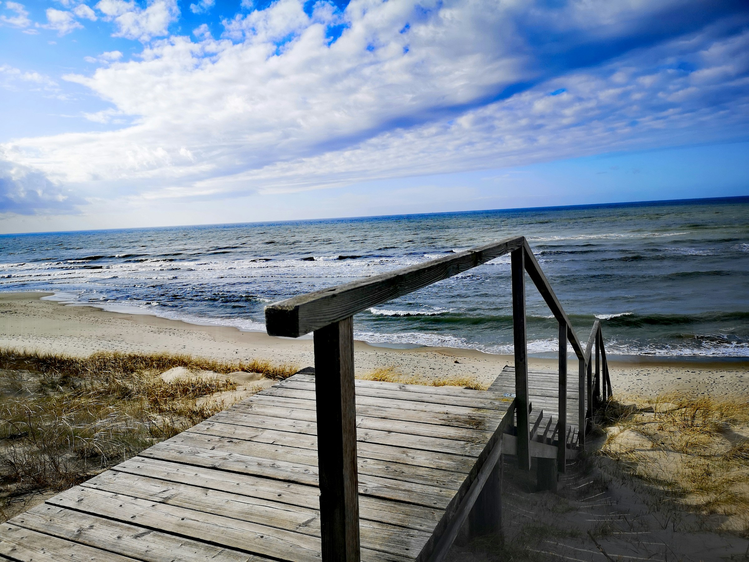 Wooden boardwalk leading to a scenic sandy beach with gentle ocean waves under a partly cloudy blue sky