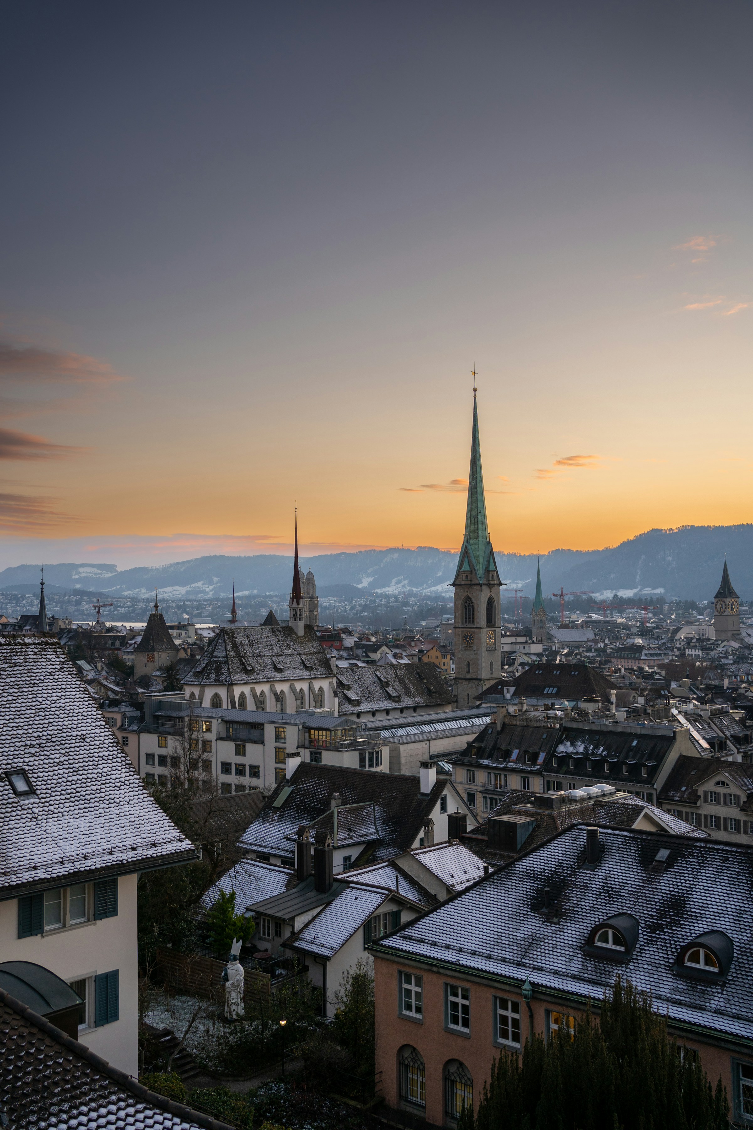 Scenic view of Zurich, Switzerland at sunset with snow-dusted rooftops and church spires against a mountainous backdrop